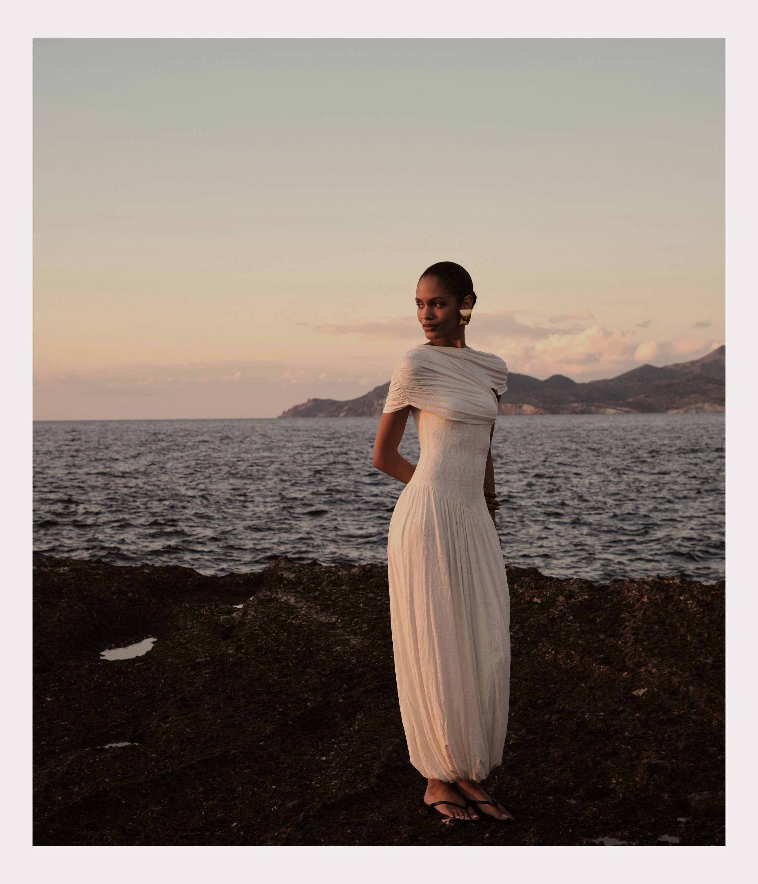 A woman in a long, elegant, cream-colored dress standing on a rocky shoreline with the ocean and mountains in the background during sunset.