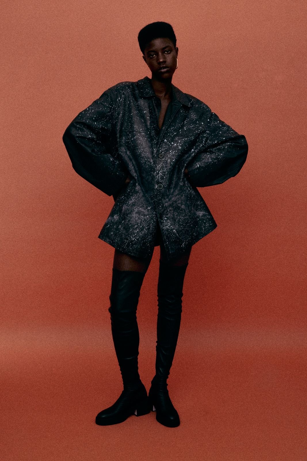 A woman wearing a dark oversized jacket with a galaxy print, black thigh-high boots, and shorts, standing against a solid orange background.