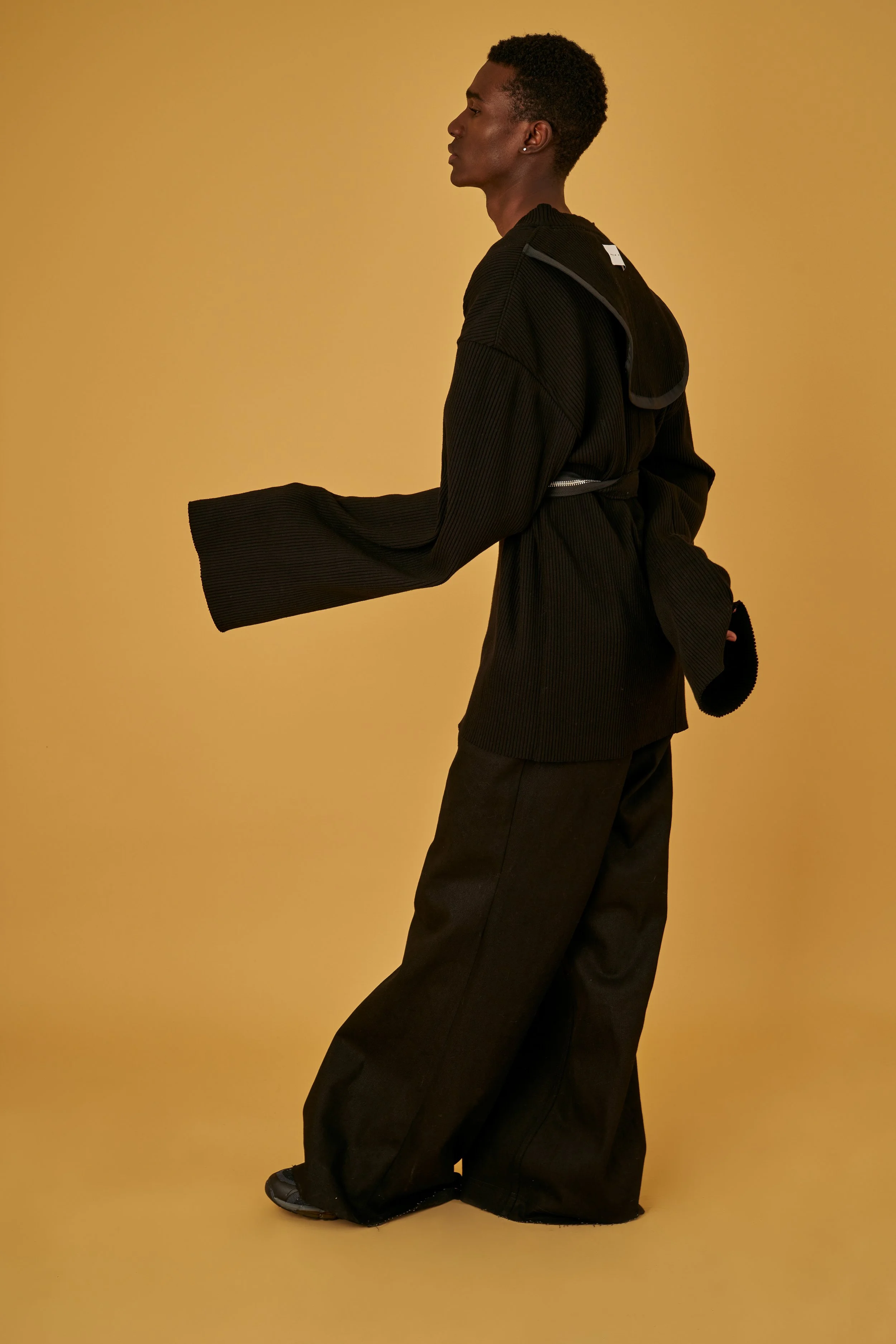 A person dressed in black, standing sideways against a yellow background, wearing oversized wide-leg pants and a loose top with extended sleeves.