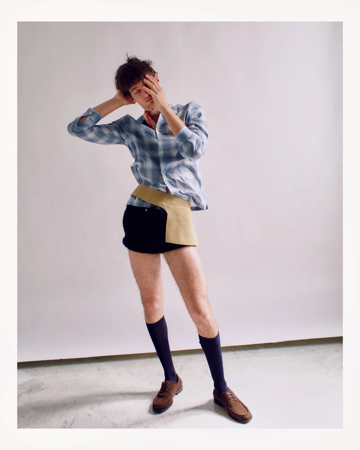 A person standing with one hand on their forehead and the other behind their head, wearing a plaid shirt, a unique skirt, dark socks, and brown shoes, standing against a plain white background.