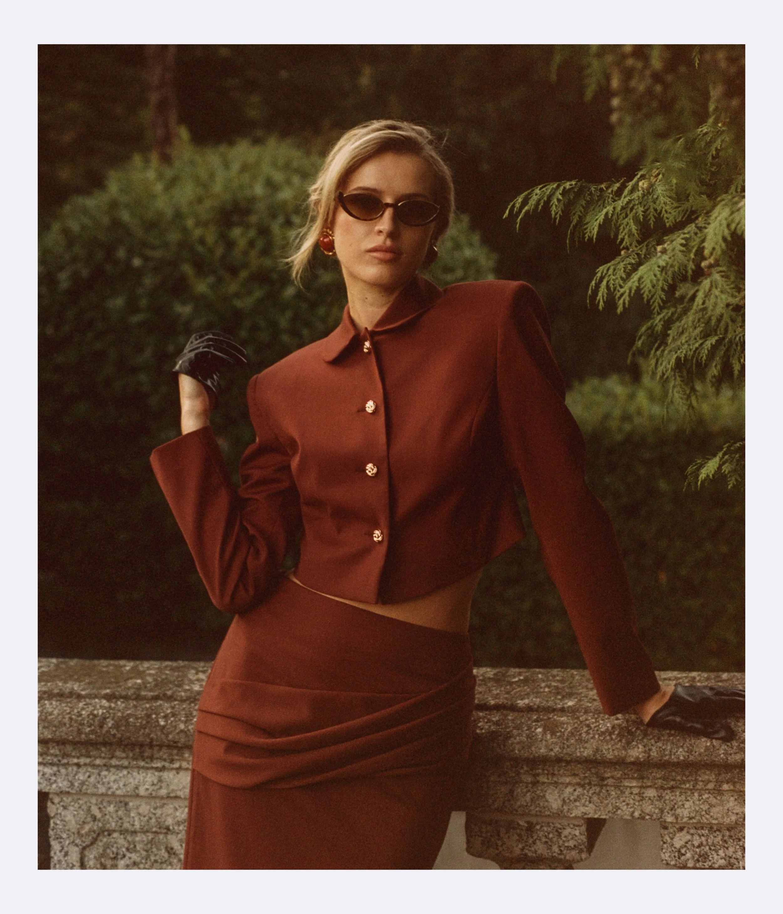 A woman dressed in a brown cropped blazer and matching skirt, wearing sunglasses and gloves, stands outdoors leaning on a stone railing with greenery in the background.