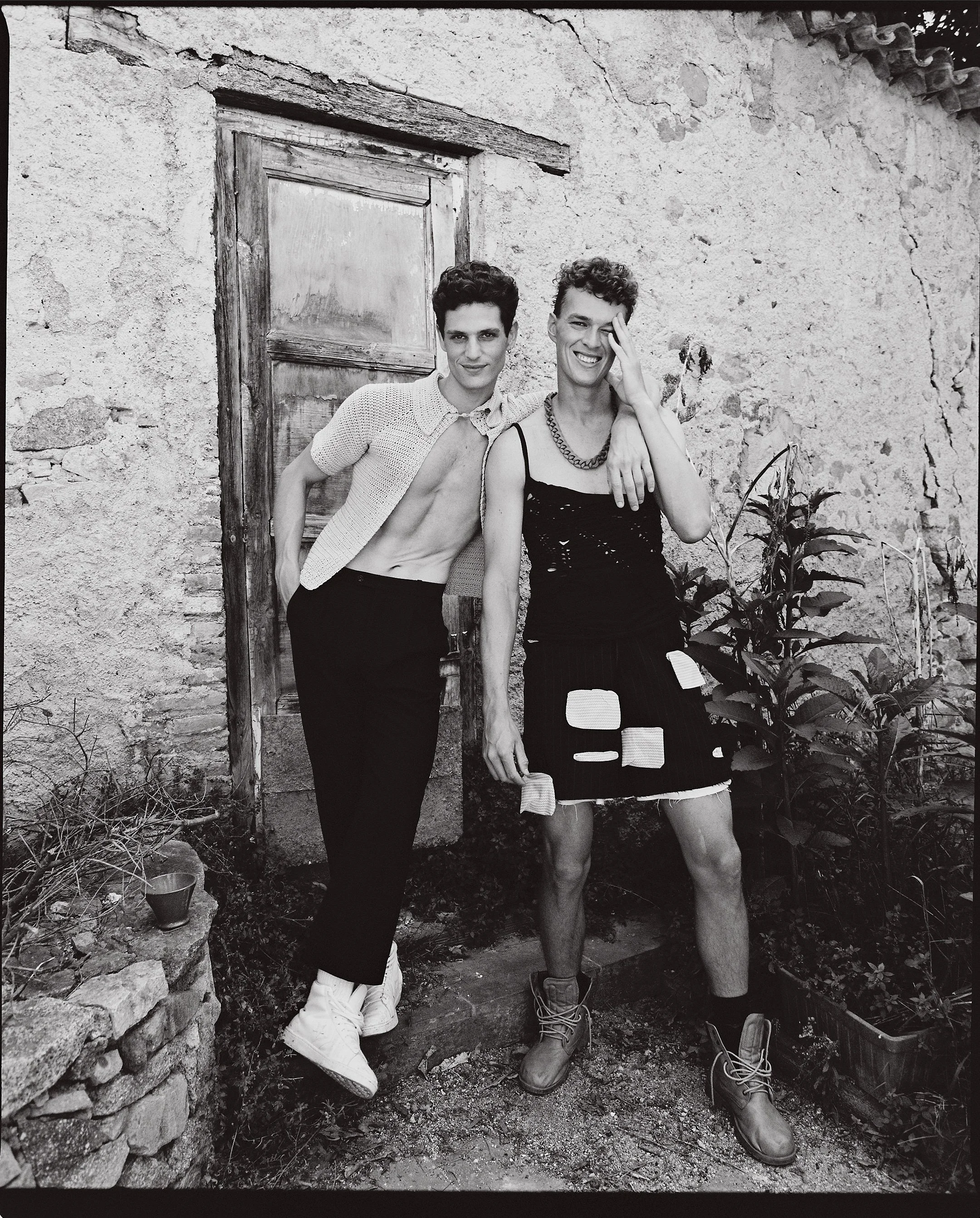 Two young men standing outside near a stone wall and plants, one with curly hair in a sleeveless dress and chain, the other with short curly hair, open shirt, and sneakers, both smiling.