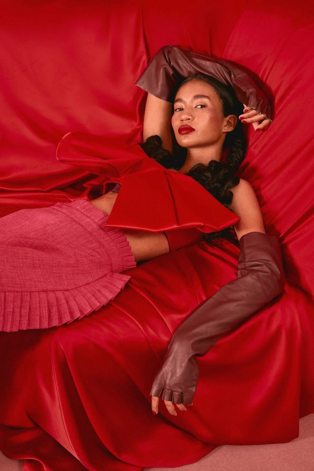 Woman lying on a red satin sofa, wearing a red dress, pink pleated skirt, and long brown leather gloves, with red lipstick and dark hair.