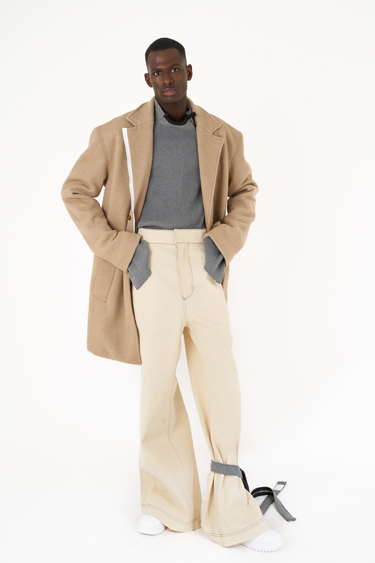 A young man with dark skin and short hair posing against a plain white background, wearing a beige coat over a gray sweater with gray cuffs, cream-colored wide-leg pants, and white sneakers with gray straps.