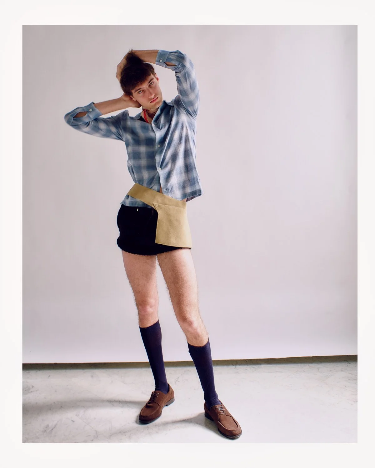A young man with messy dark hair and a serious expression wearing an unbuttoned blue plaid shirt, a beige apron, black shorts, dark knee-high socks, and brown shoes standing against a plain white background.