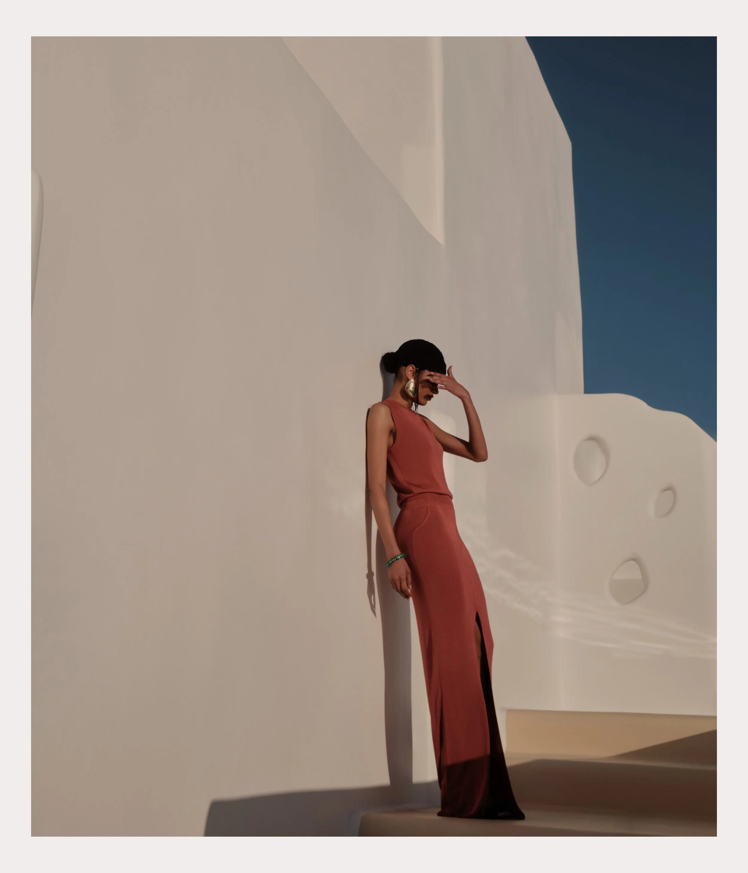 Woman in a sleeveless reddish-brown dress with a slit, standing against a white textured wall with curves, looking down with her hand on her forehead, in bright sunlight, with clear blue sky in the background.