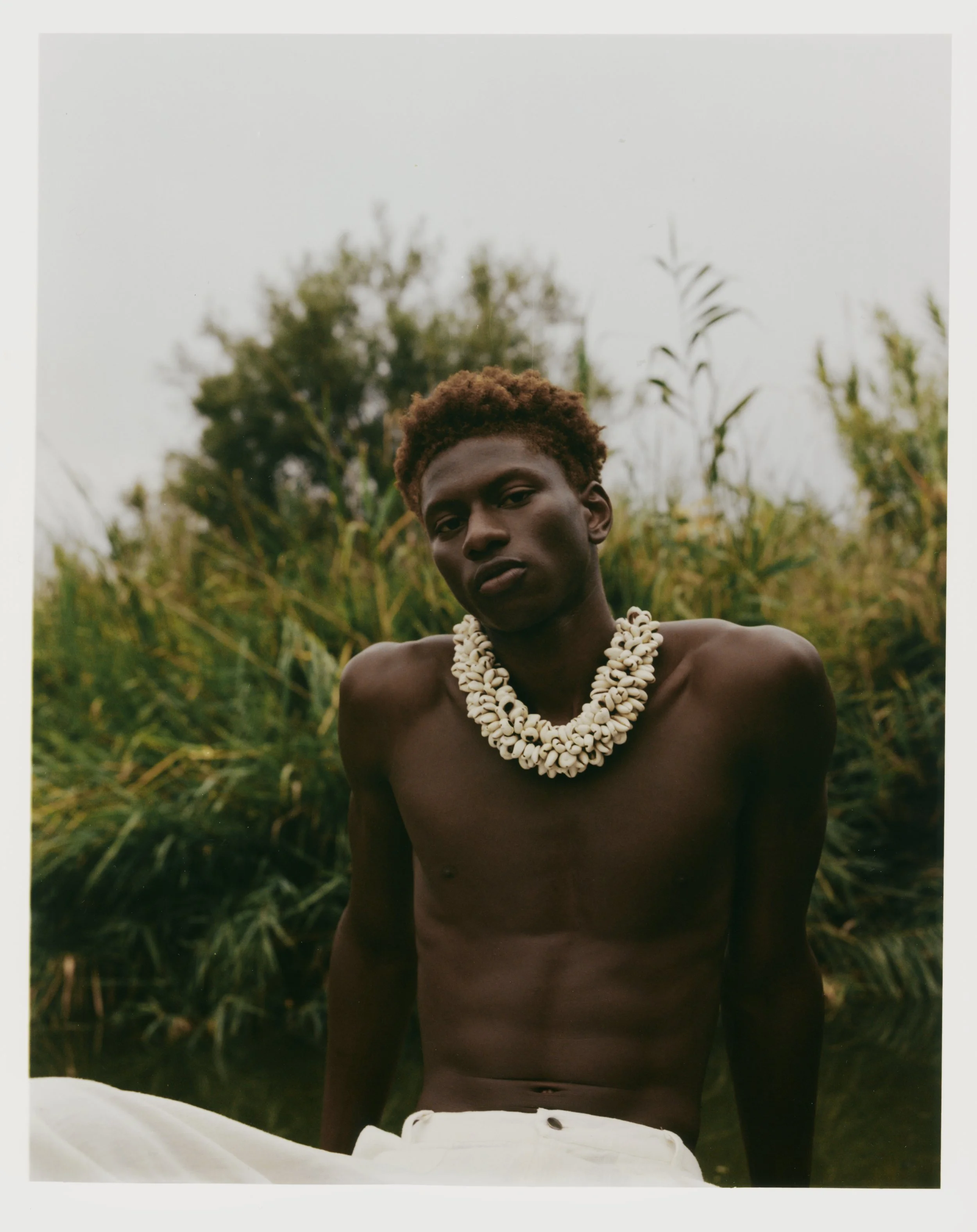 A young man with dark skin and short, curly hair wearing a large shell necklace, shirtless, outdoors with tall green plants and trees in the background.