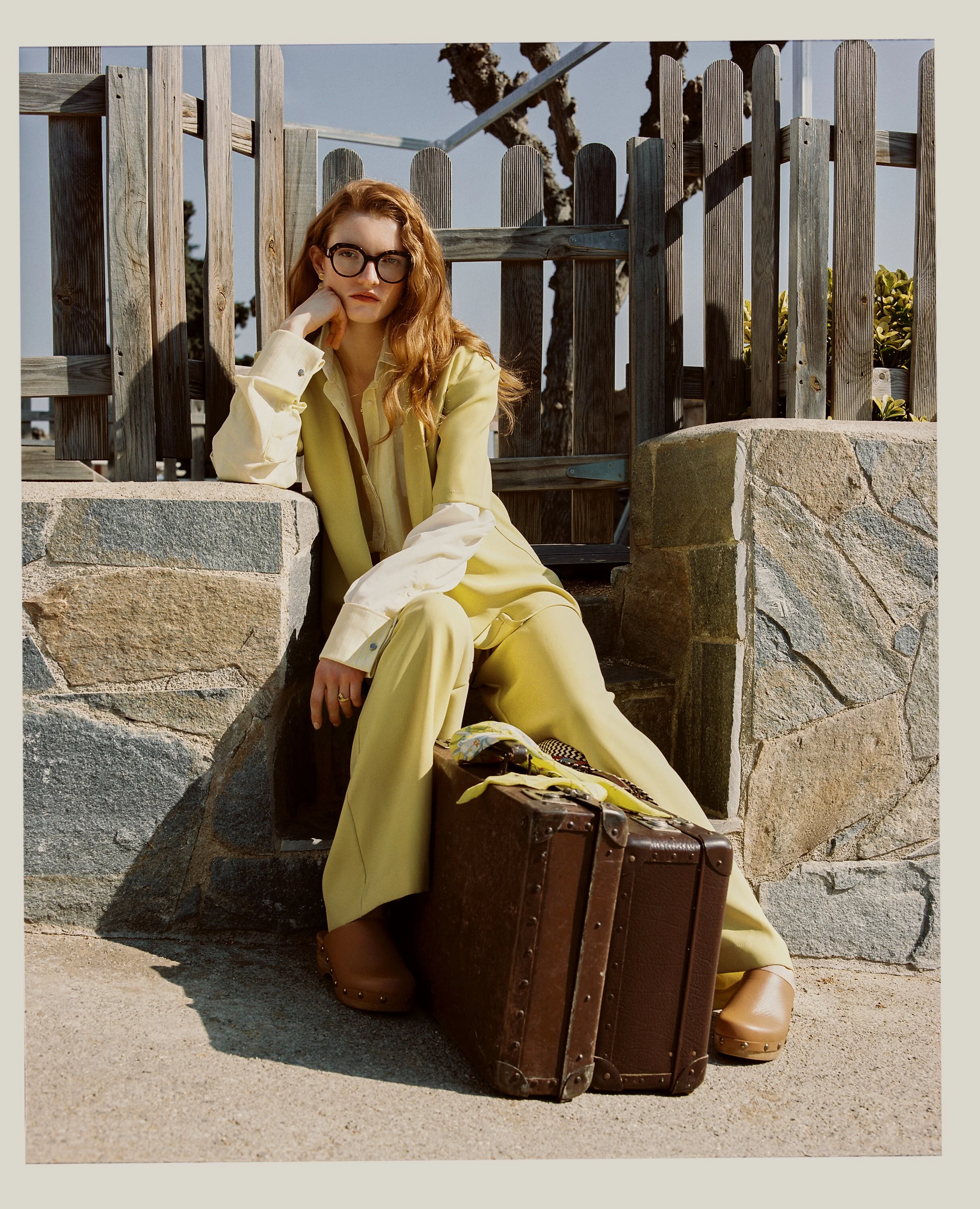 A woman with red hair, glasses, wearing a yellow outfit, sitting on stone stairs next to a vintage suitcase, outdoors near a wooden fence.