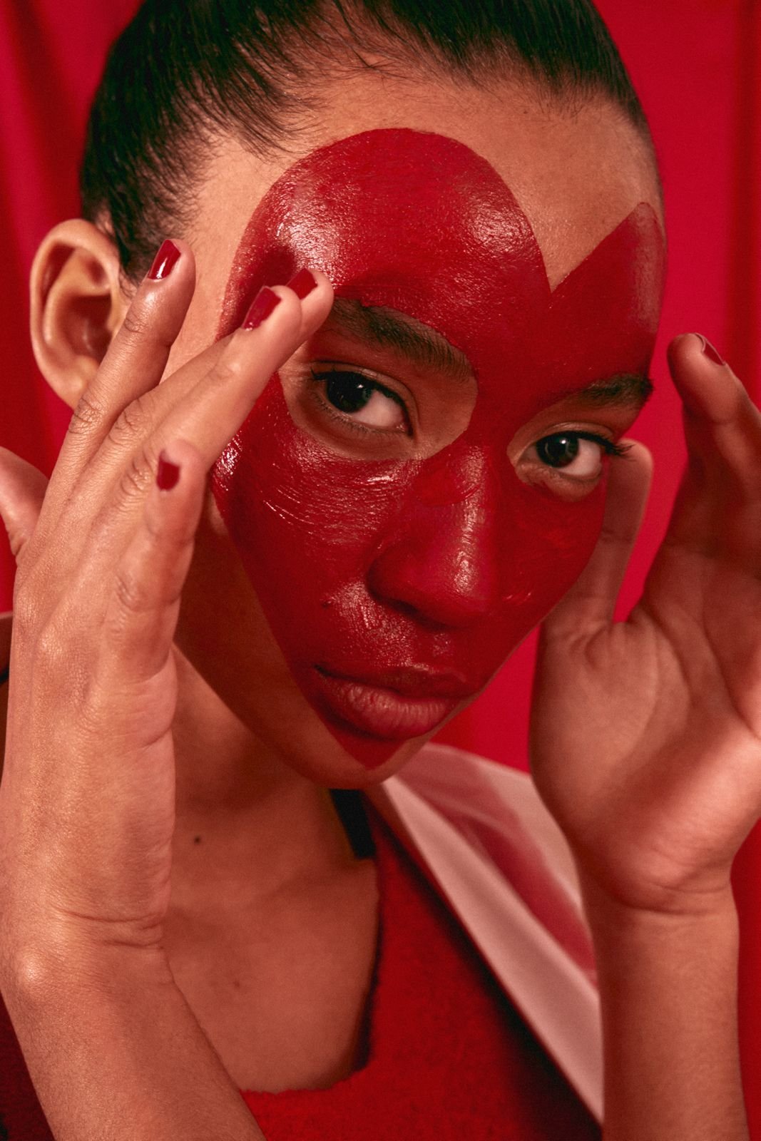 A woman with a red painted face and red lipstick, holding her hands near her face with red painted nails, against a red background.