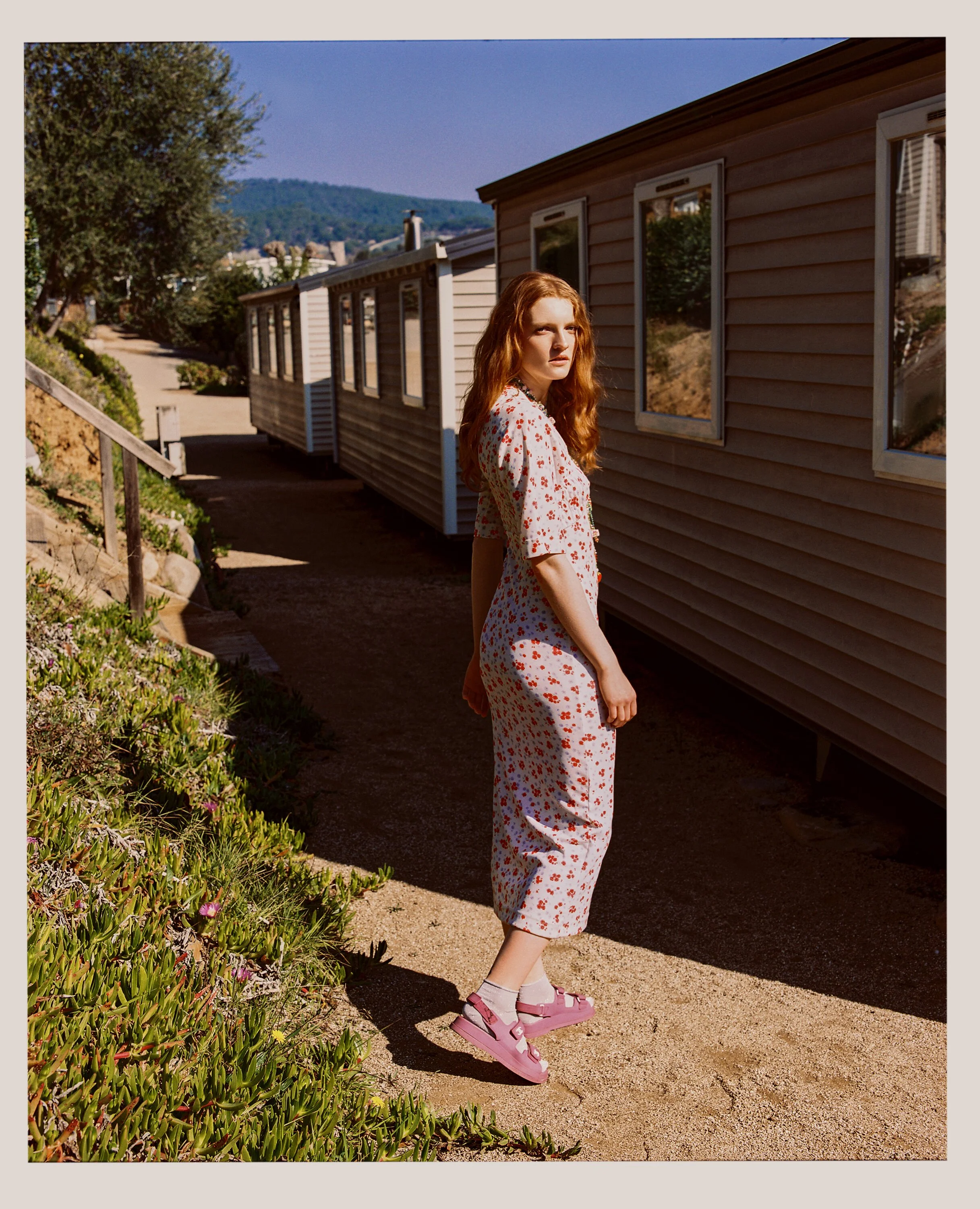 A young woman with long red hair stands on a sandy path beside a tan house with white-framed windows, wearing a matching floral pajama set, pink sandals, and socks, gazing into the distance with a serious expression, with trees and hills in the backg