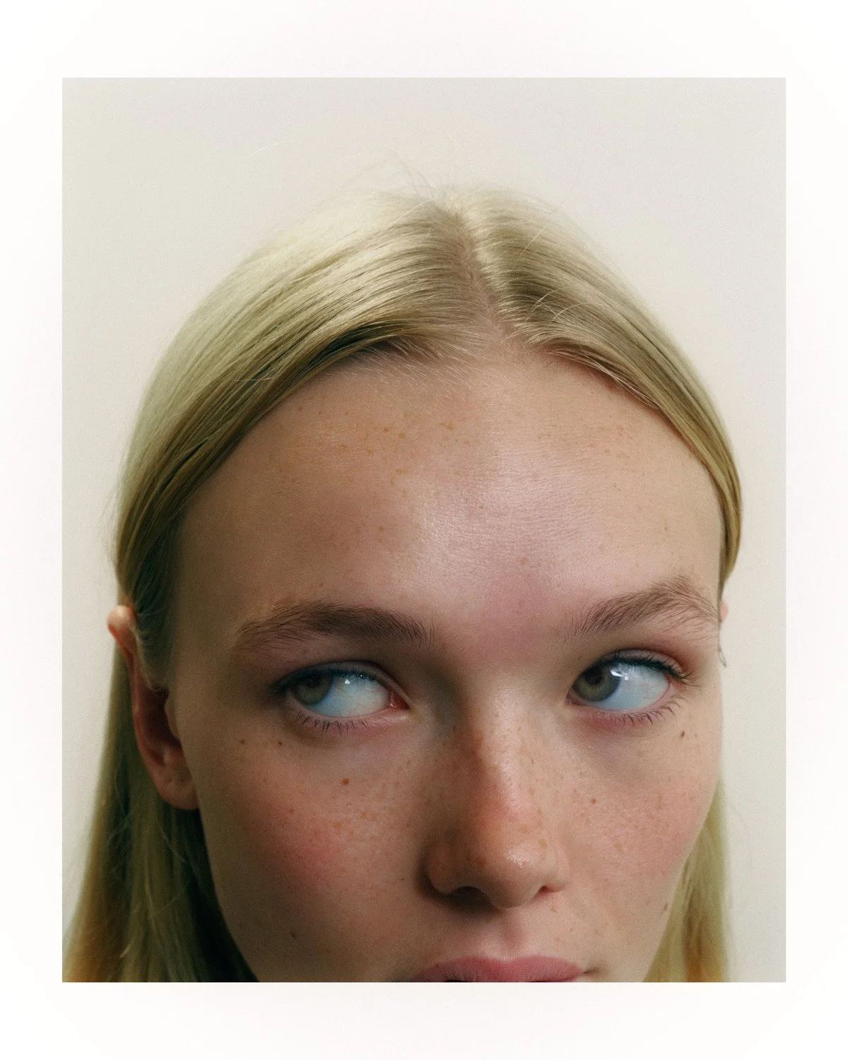 Close-up of a young woman's face with blonde hair, light skin, freckles, and blue eyes, looking slightly upward and to the side against a plain white background.