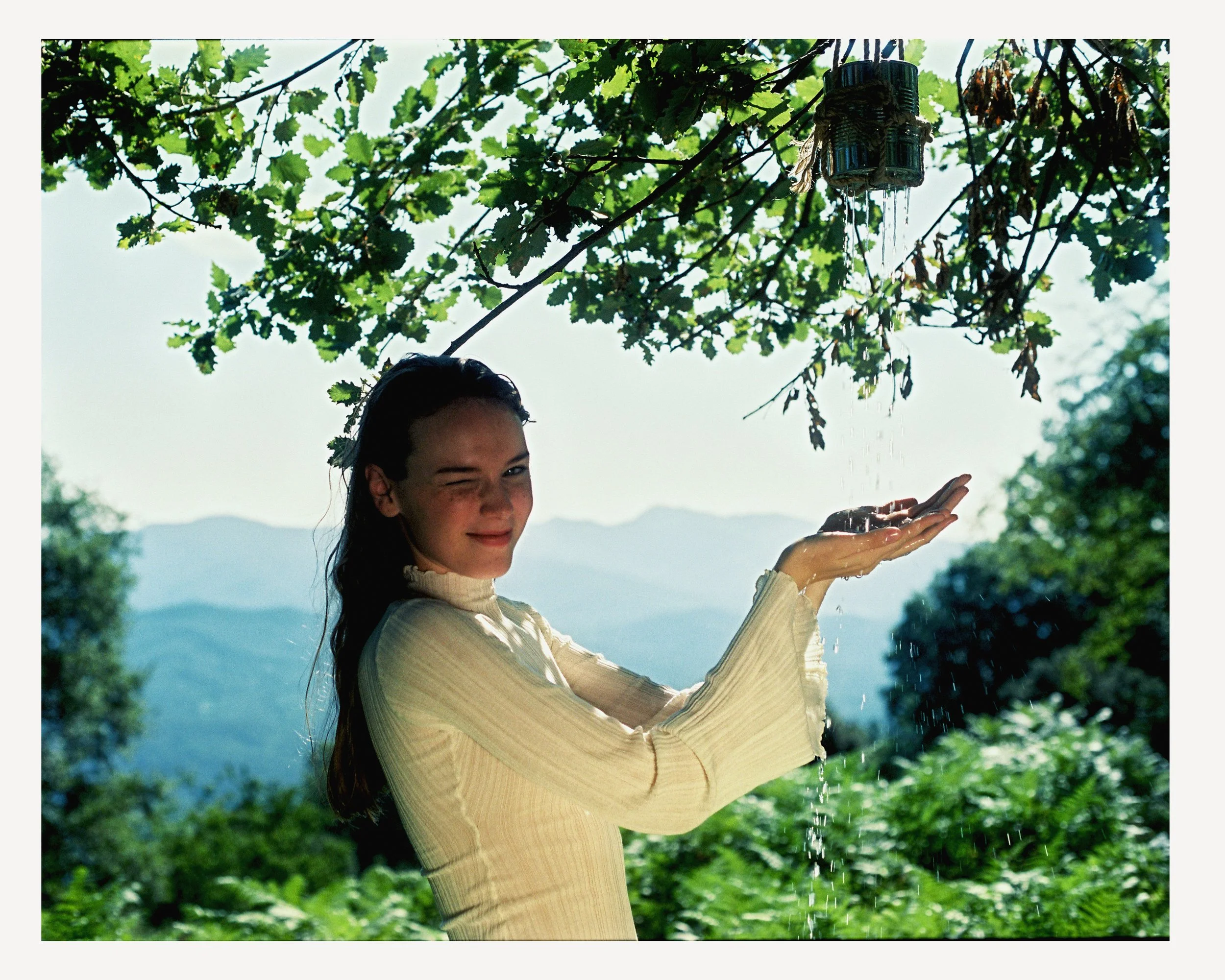 A woman outdoors in a scenic natural setting with mountains in the background. She is wearing a light-colored long-sleeve top and is smiling and winking while holding her hands under a water stream from a makeshift rain collector hanging from a tree 