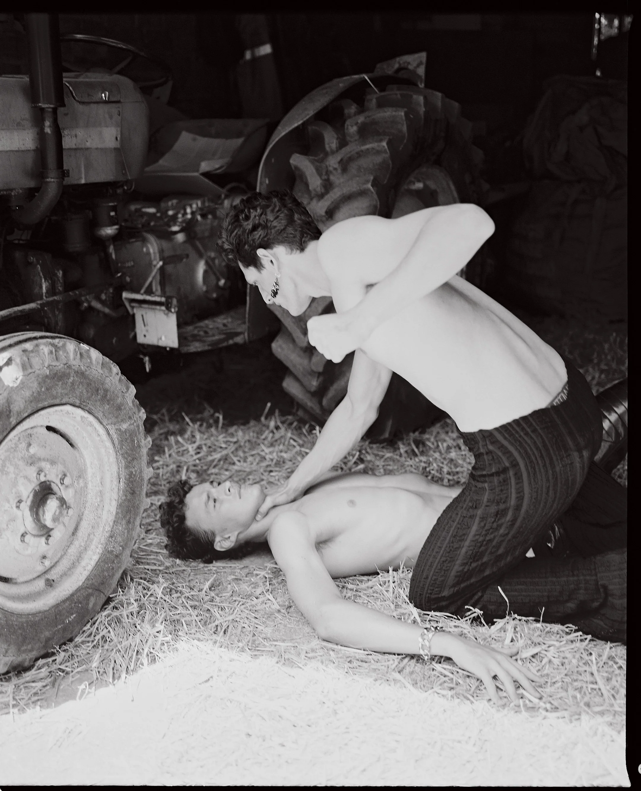 A black and white photo of a woman lying on the ground shirtless with a man kneeling over her, appearing to be performing CPR or a similar emergency procedure, next to a large tractor tire in a barn or garage setting.