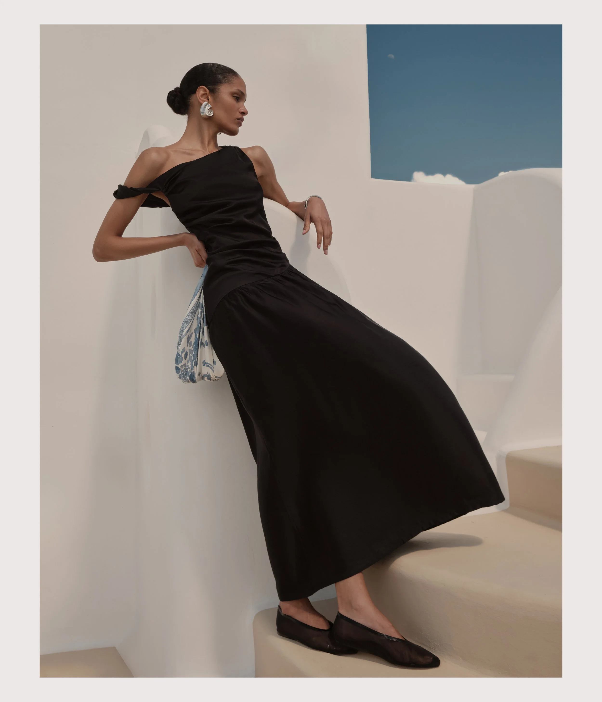 A woman in a black dress and black flats leaning against a white wall, with a blue sky visible through an opening behind her.