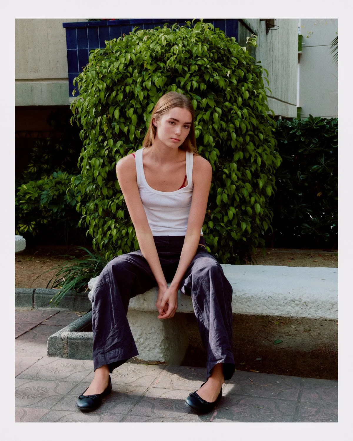 A young woman with light skin and long blonde hair sits on a concrete bench outdoors, wearing a white tank top, black wide-leg pants, and black ballet flats, with her hands resting between her knees, in front of a green bush and a building with beige