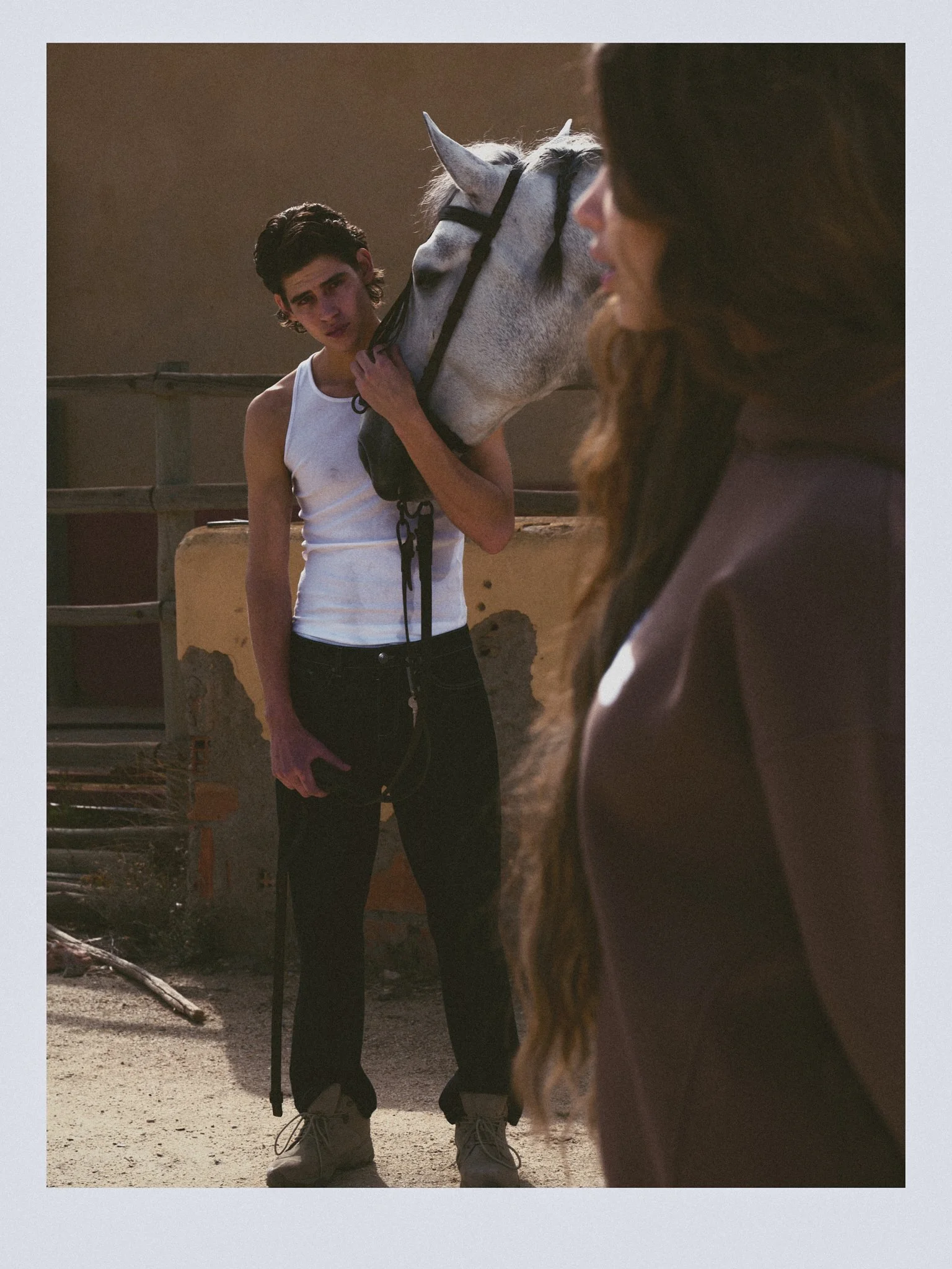 A young man with dark hair wearing a white tank top and black pants, holding a white horse's bridle, standing outdoors with a brown fence behind him. A woman with long hair, partially in view, is in the foreground.