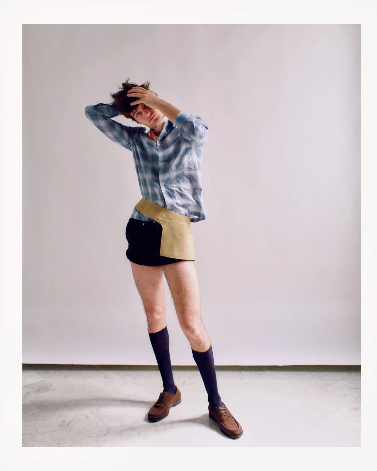 A person standing in front of a plain white wall, wearing a plaid shirt, a beige and black skirt, black socks, and brown shoes, with messy hair and one hand on their head.