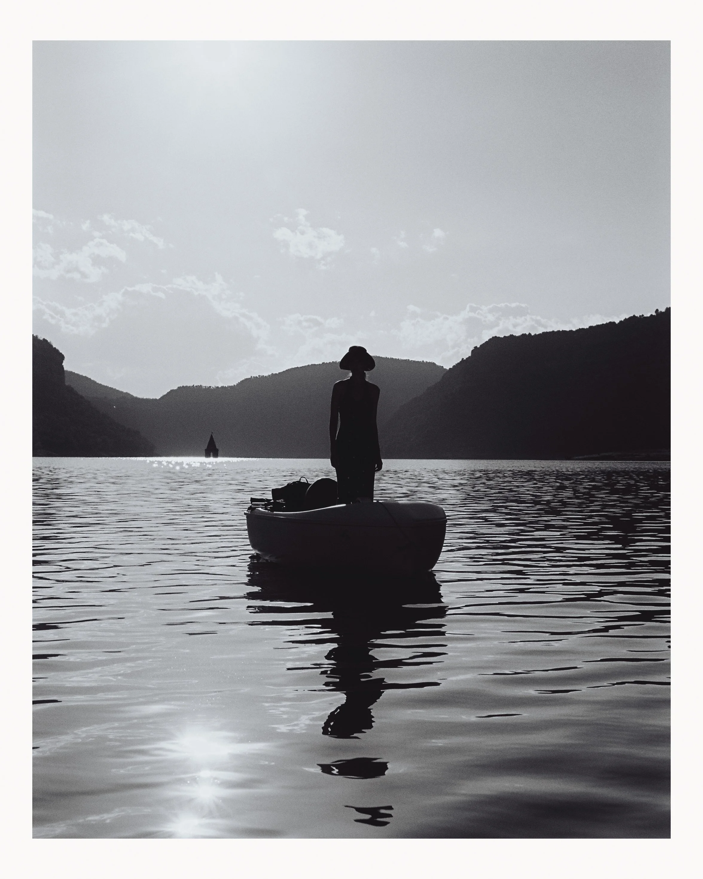 Silhouette of a woman standing in a boat on a calm body of water, with mountains in the background and a small boat or structure far in the distance, under a partly cloudy sky.