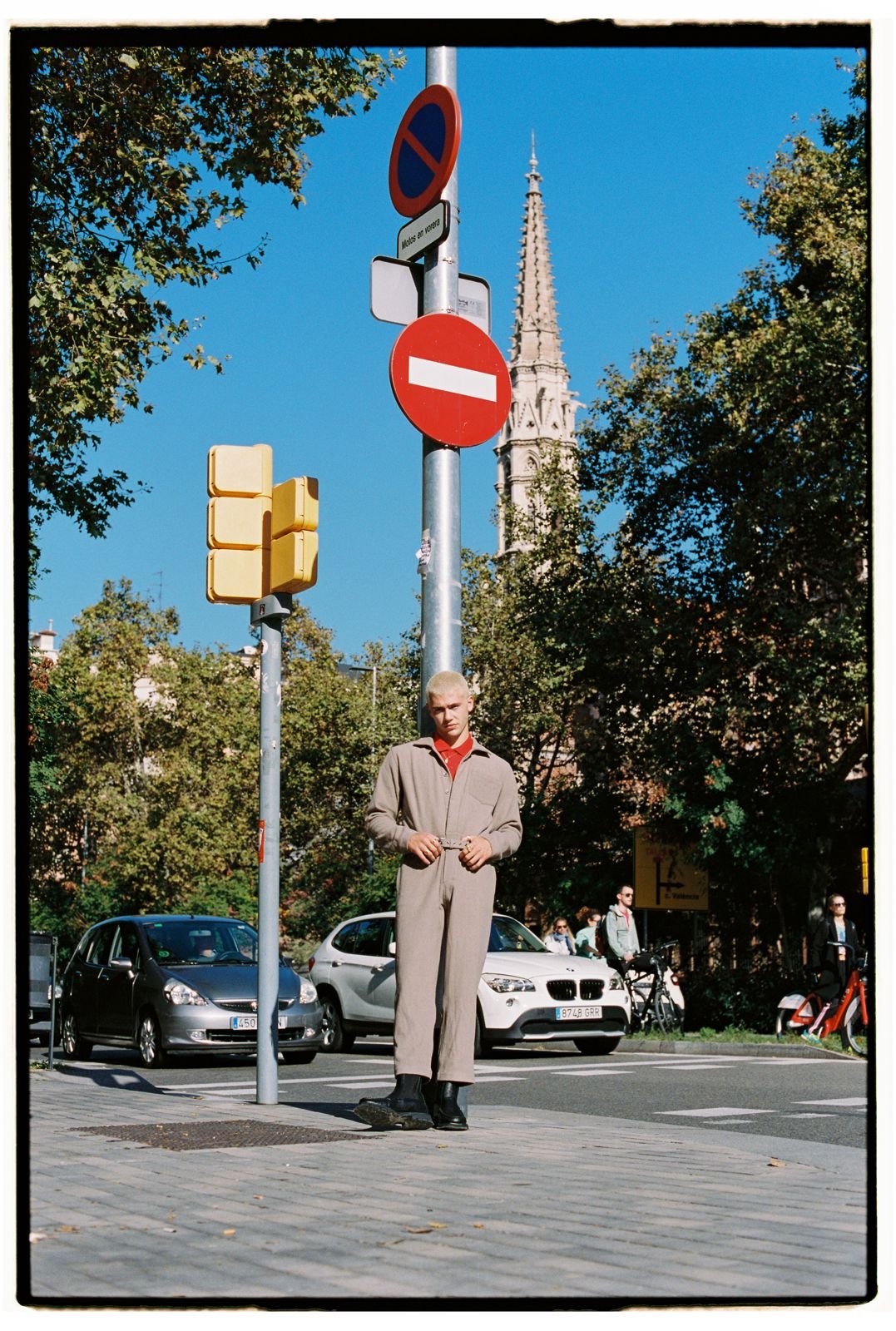 Young man with a buzz cut wearing a beige suit and black shoes standing in front of traffic signs on a city street, with cars, people, trees, and a church spire in the background on a sunny day.