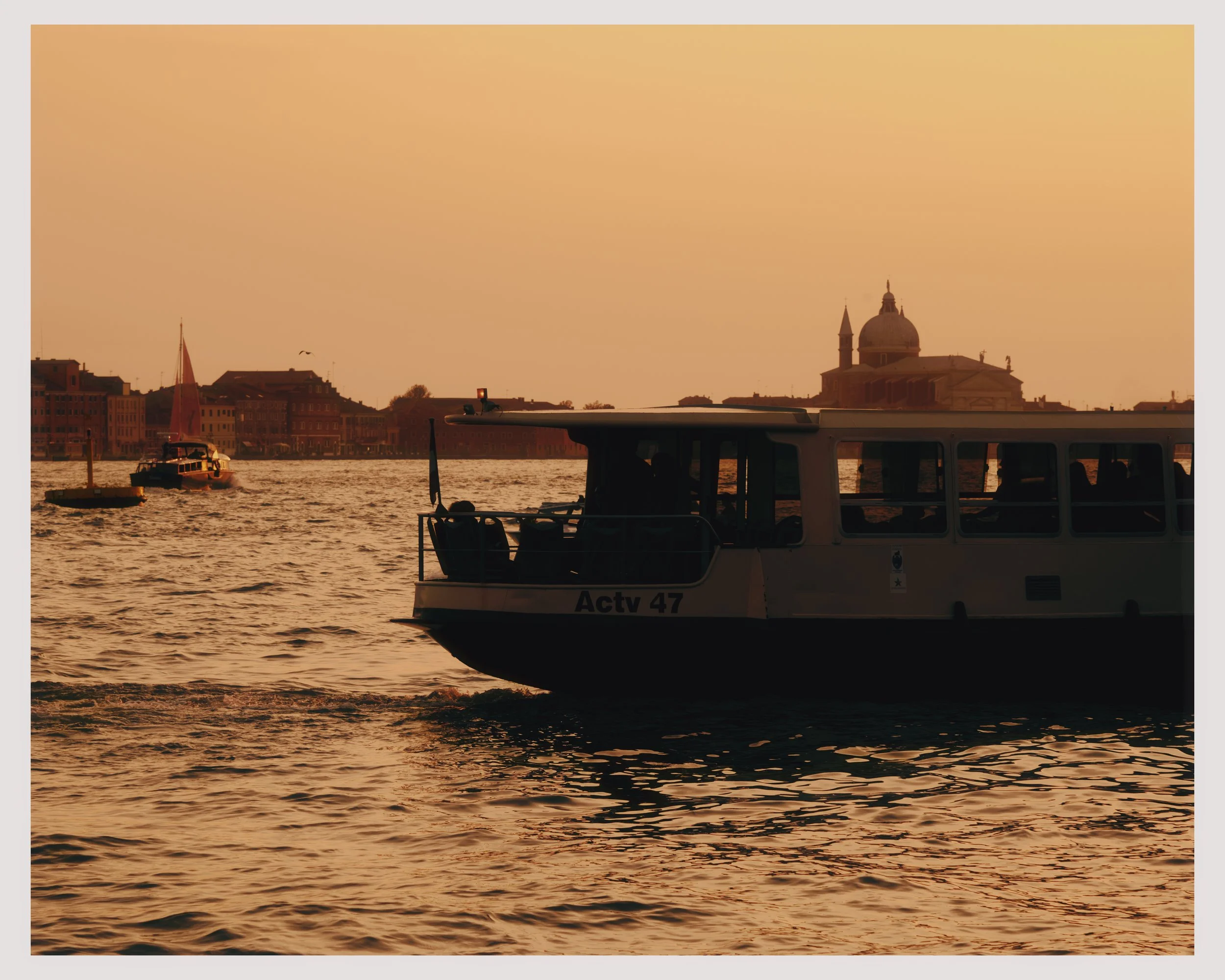 A boat labeled 'Actv 47' sailing on a body of water during sunset, with a city skyline and a church with a dome and a tower in the background.