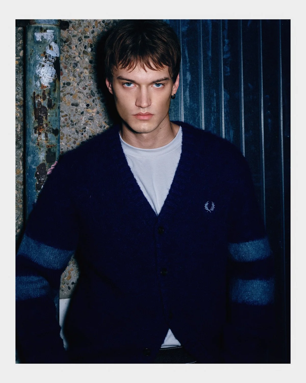 Young man with brown hair and blue eyes wearing a dark blue cardigan with gray stripes on the sleeves, standing against a textured wall with a blue panel.