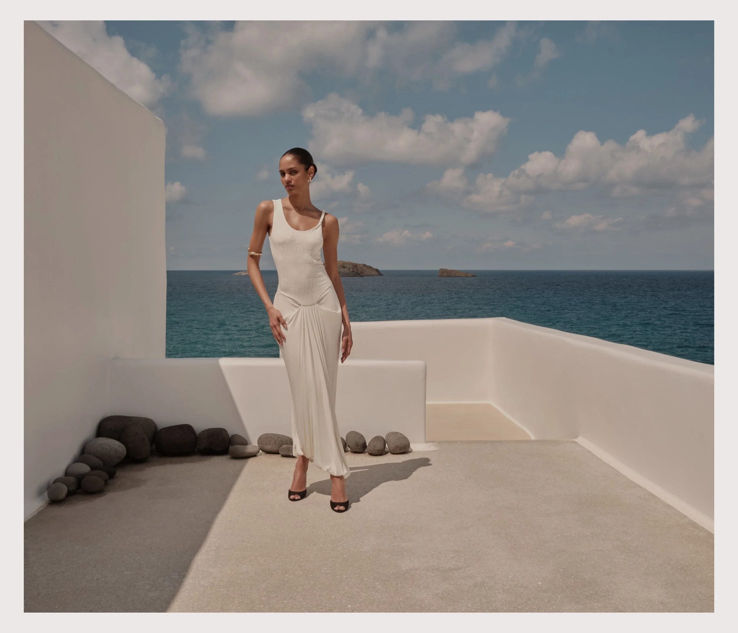 A woman in a white dress and black high heels stands on a terrace by the ocean. The sky is partly cloudy, and there are small islands in the distance. The terrace has white walls, a stone decoration, and a smooth beige floor.