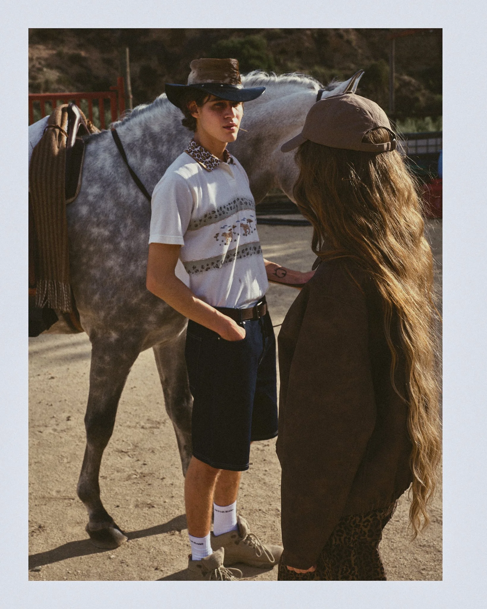Two young adults talking outdoors, with a white horse in the background. One wears a wide-brimmed hat, graphic t-shirt, shorts, and sneakers. The other has long hair, a cap, and dark clothing.