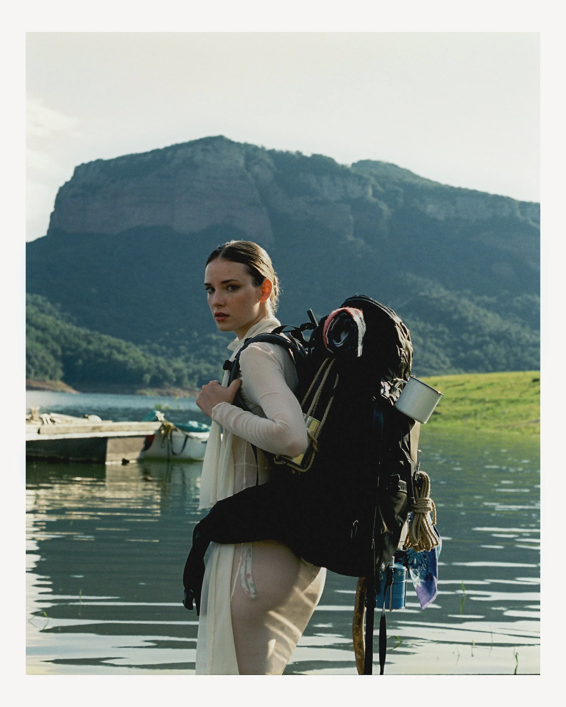 A woman with a backpack standing by a body of water with mountains in the background.