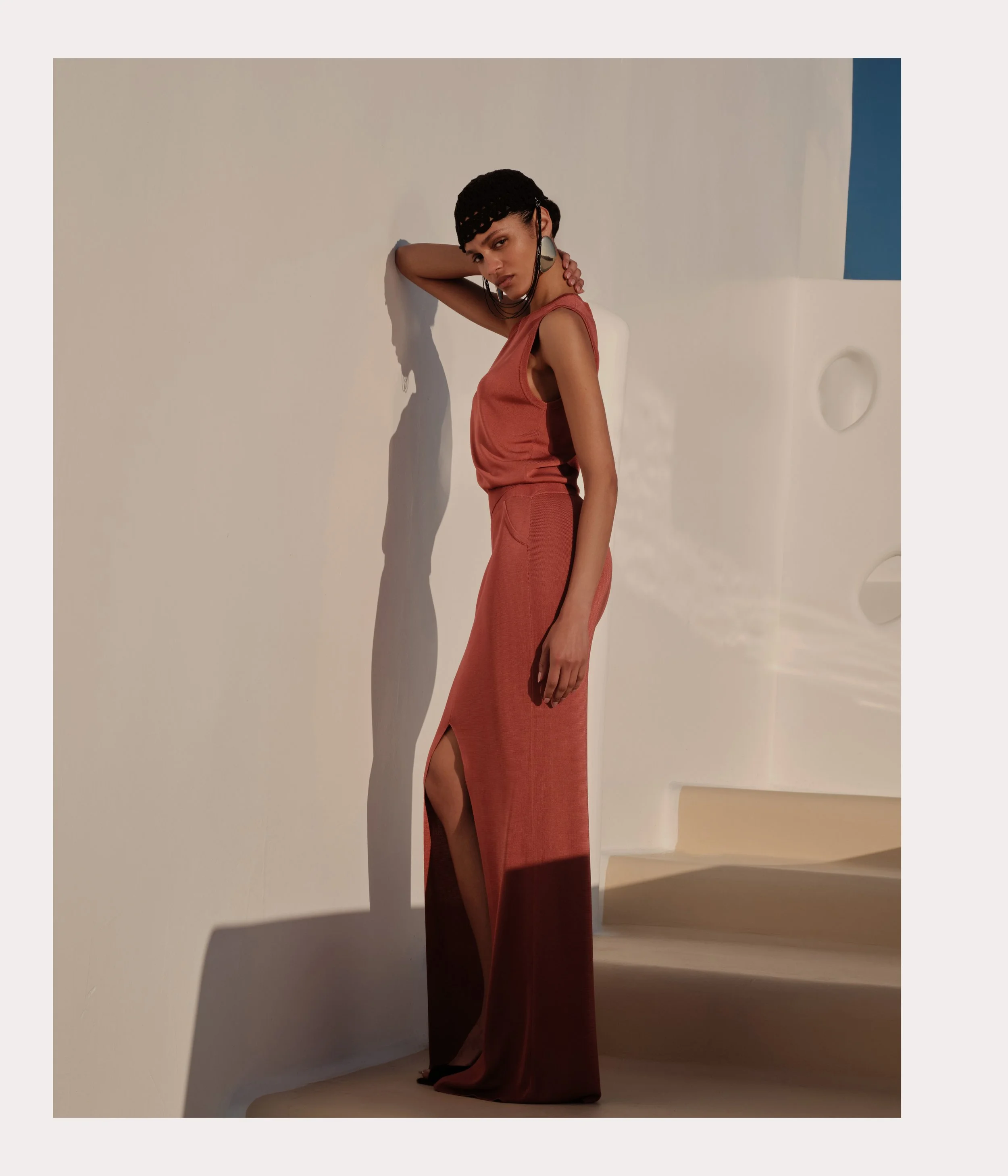 A woman wearing a sleeveless, rust-colored dress with a slit, standing against a white wall with shadows, in a minimalist setting with curved stairs and circular wall accents, under warm lighting.