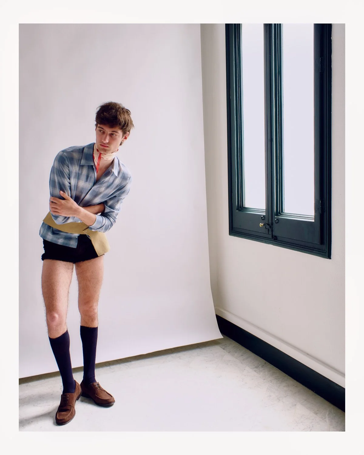 Young man wearing a plaid shirt, shorts, and knee-high socks standing in a photography studio with a white backdrop and black-framed window, with no underwear.