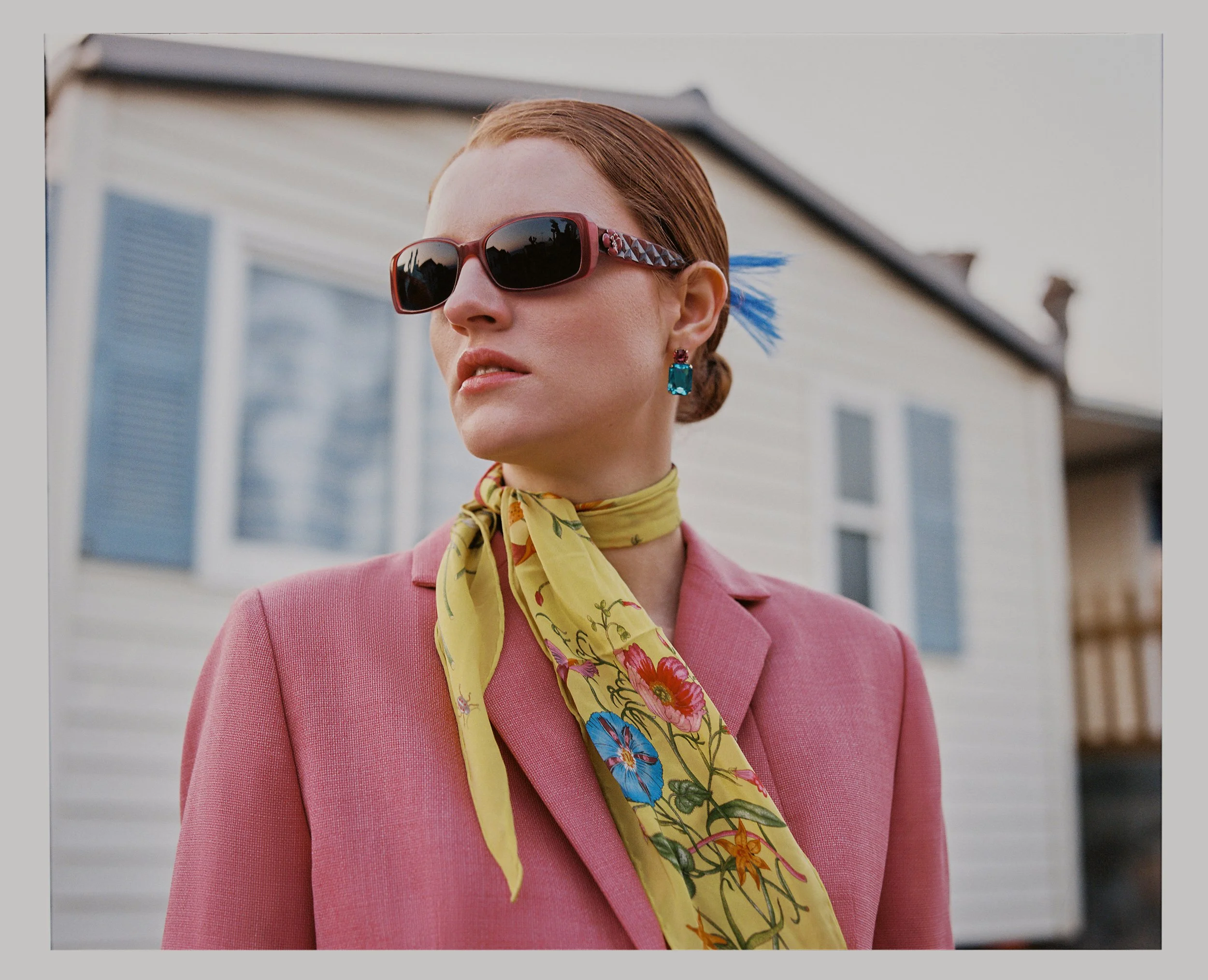 A woman with styled red hair wearing large sunglasses, turquoise earrings, a yellow floral scarf, and a pink blazer stands outside in front of a house with blue shutters.