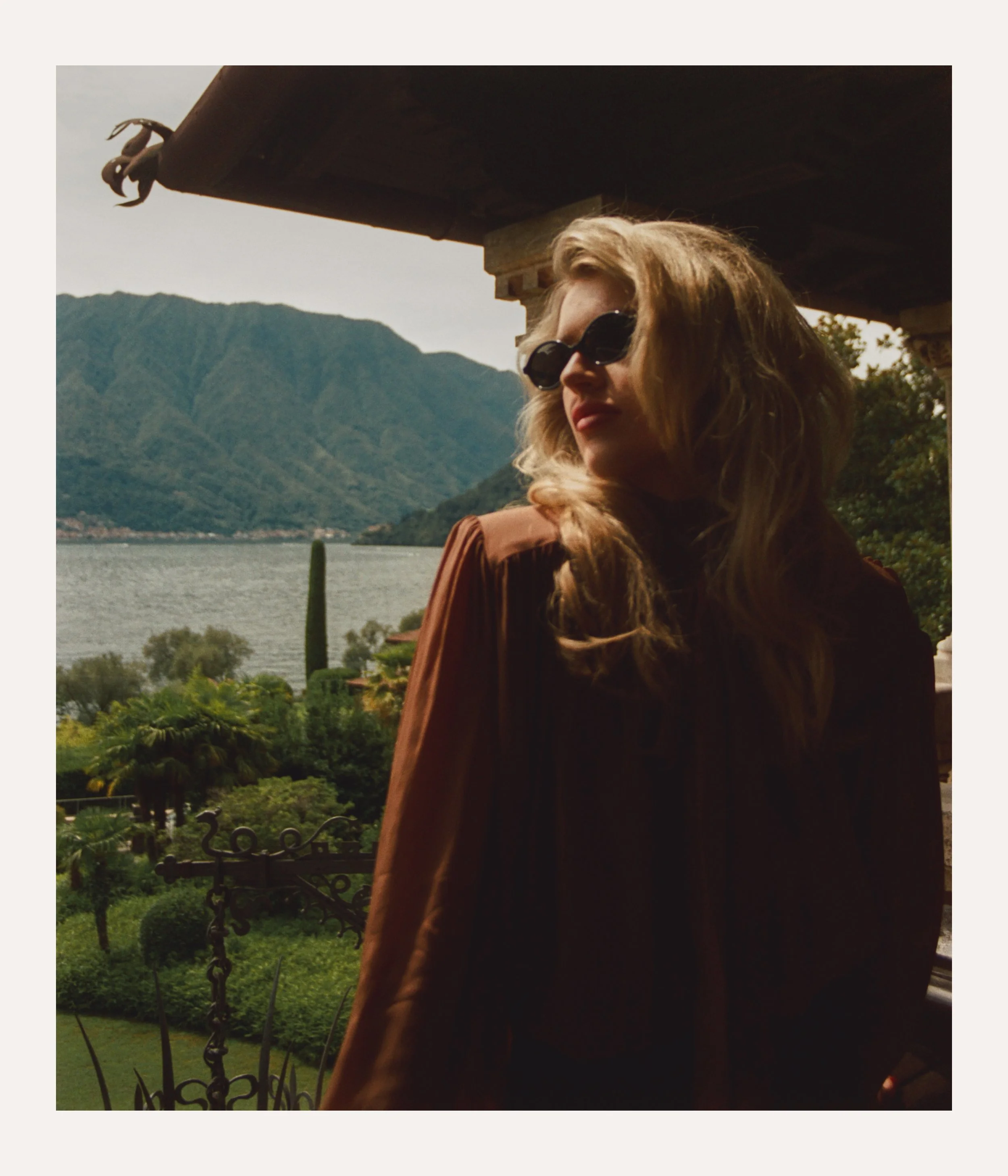 A woman with long blonde hair wearing sunglasses and a brown jacket stands on a balcony overlooking a lake and mountains.