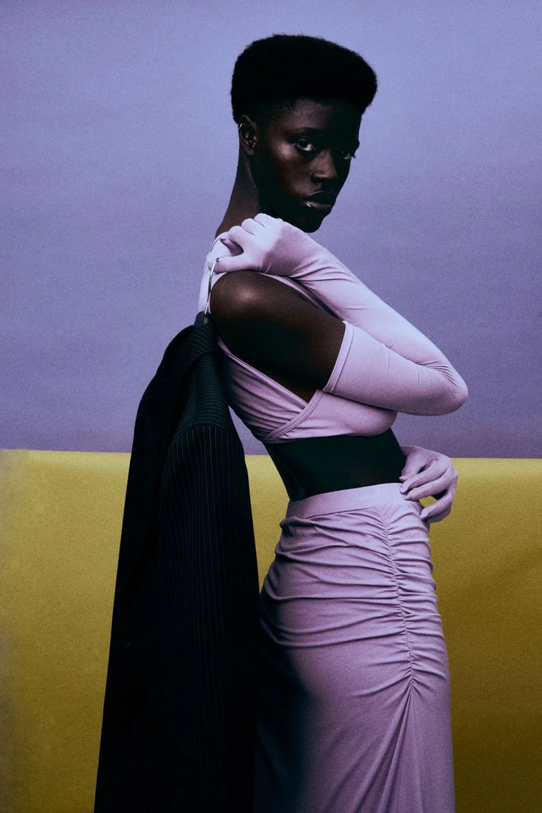 A dark-skinned woman wearing a pink body-hugging dress with a ruched skirt and long gloves, holding a black blazer over her shoulder, standing against a two-tone background of purple and yellow.