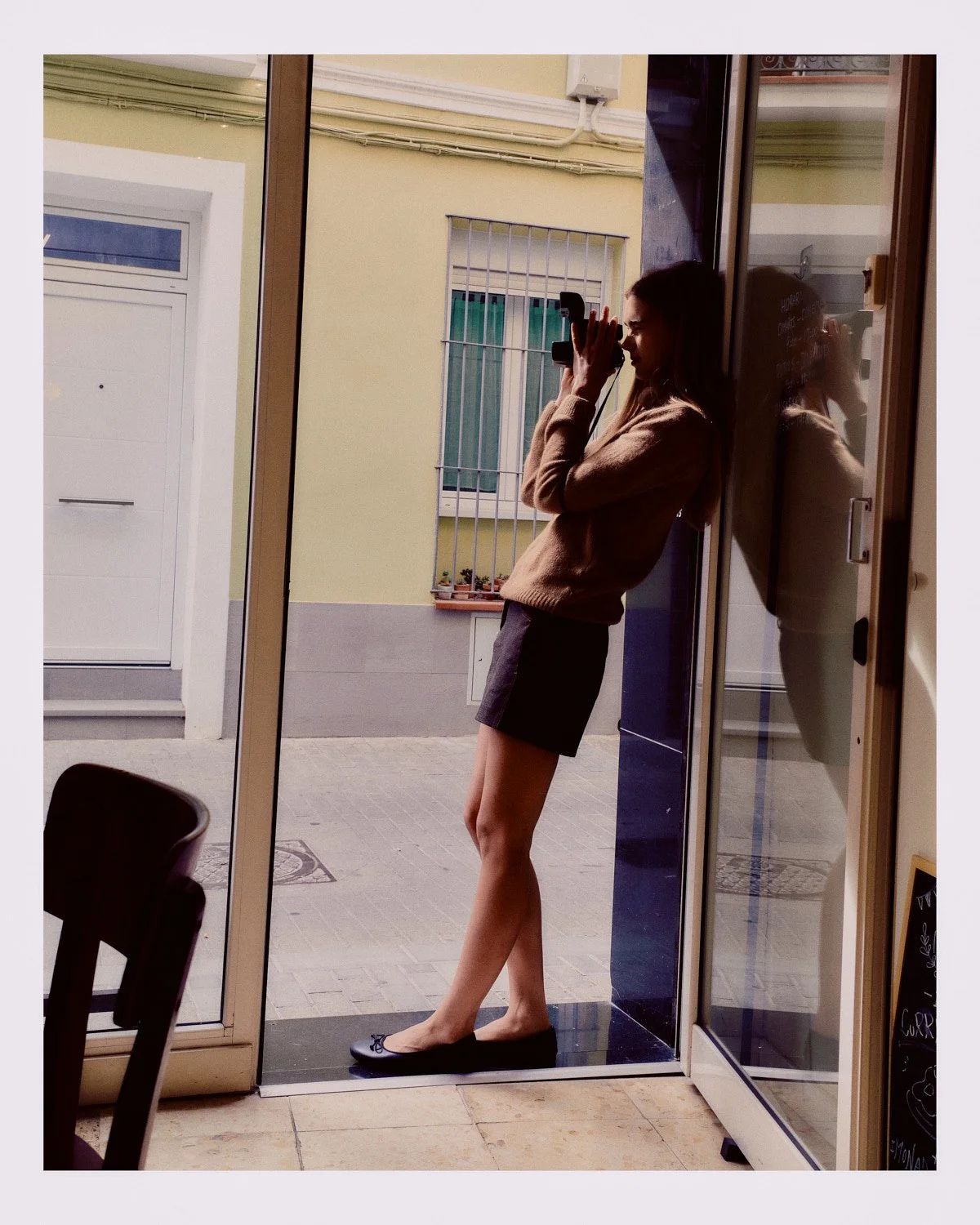 A woman standing barefoot on a black doormat, taking a photo with a small camera while leaning against a glass door frame, with her reflection visible in the glass.