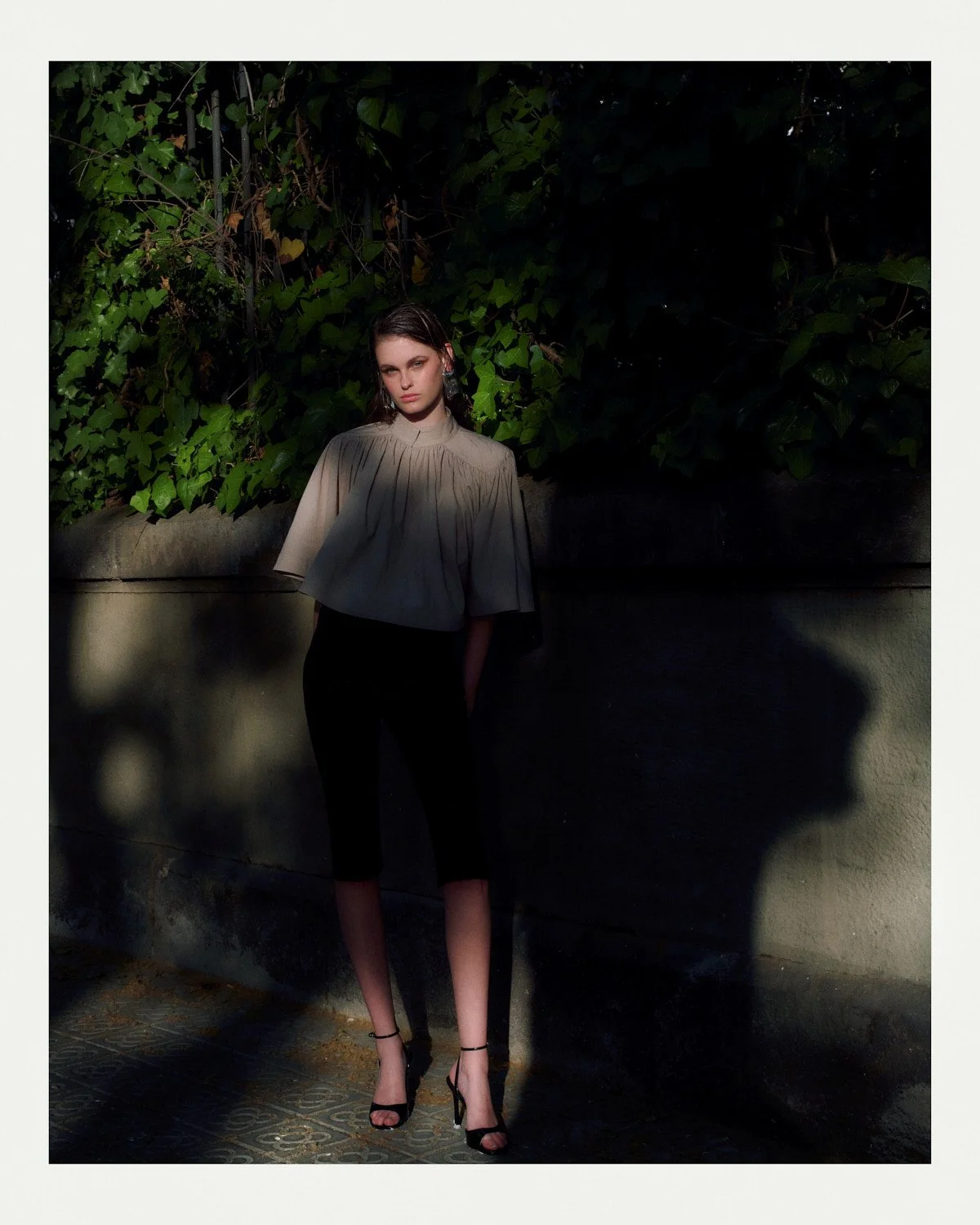 A woman with dark hair and a beige top with wide sleeves stands outdoors against a dark background of greenery and a concrete wall, shadows cast on the wall.