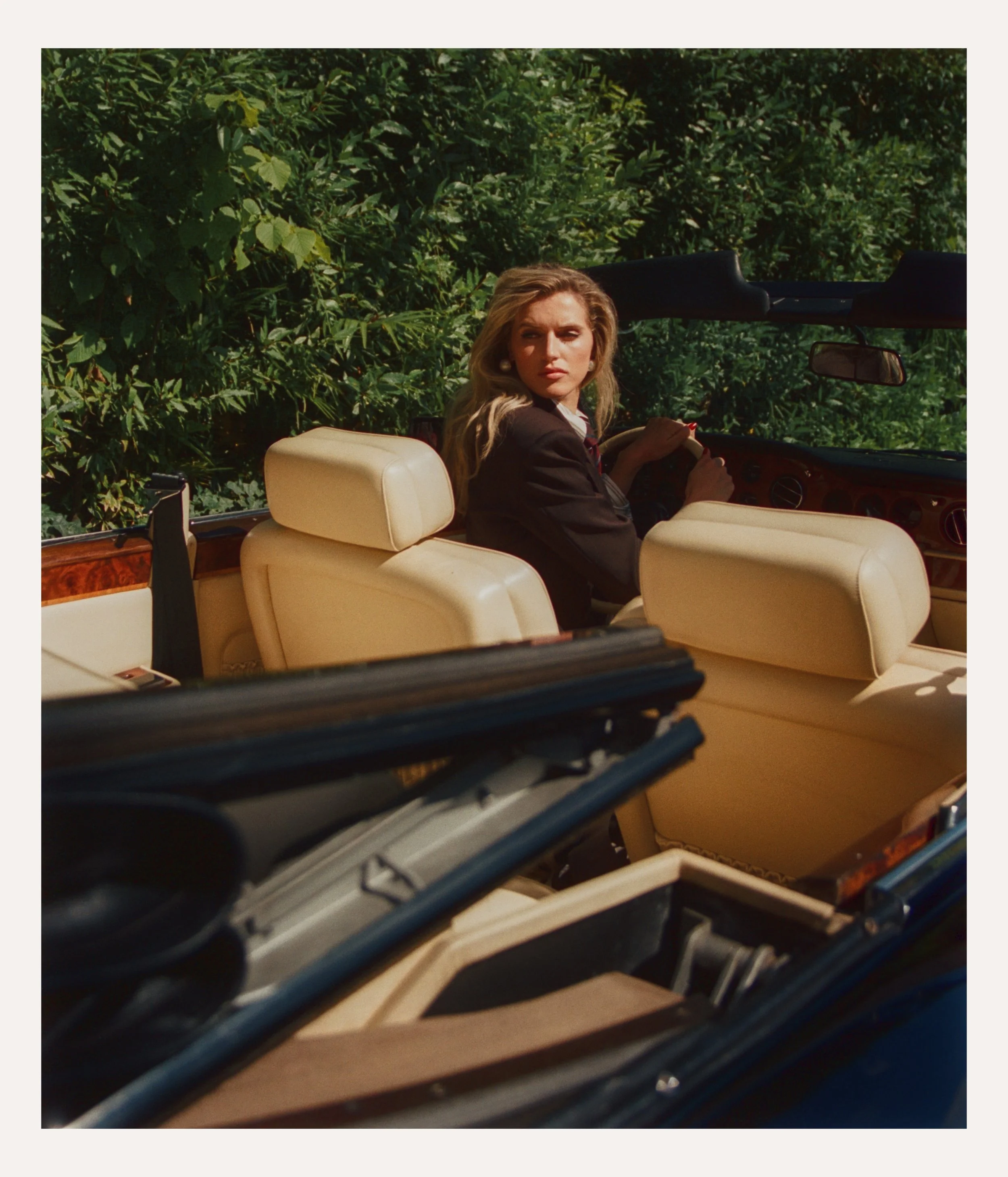 A woman with blonde hair and a black jacket sitting in the driver's seat of a vintage convertible car, looking over her shoulder with a serious expression, surrounded by lush greenery.