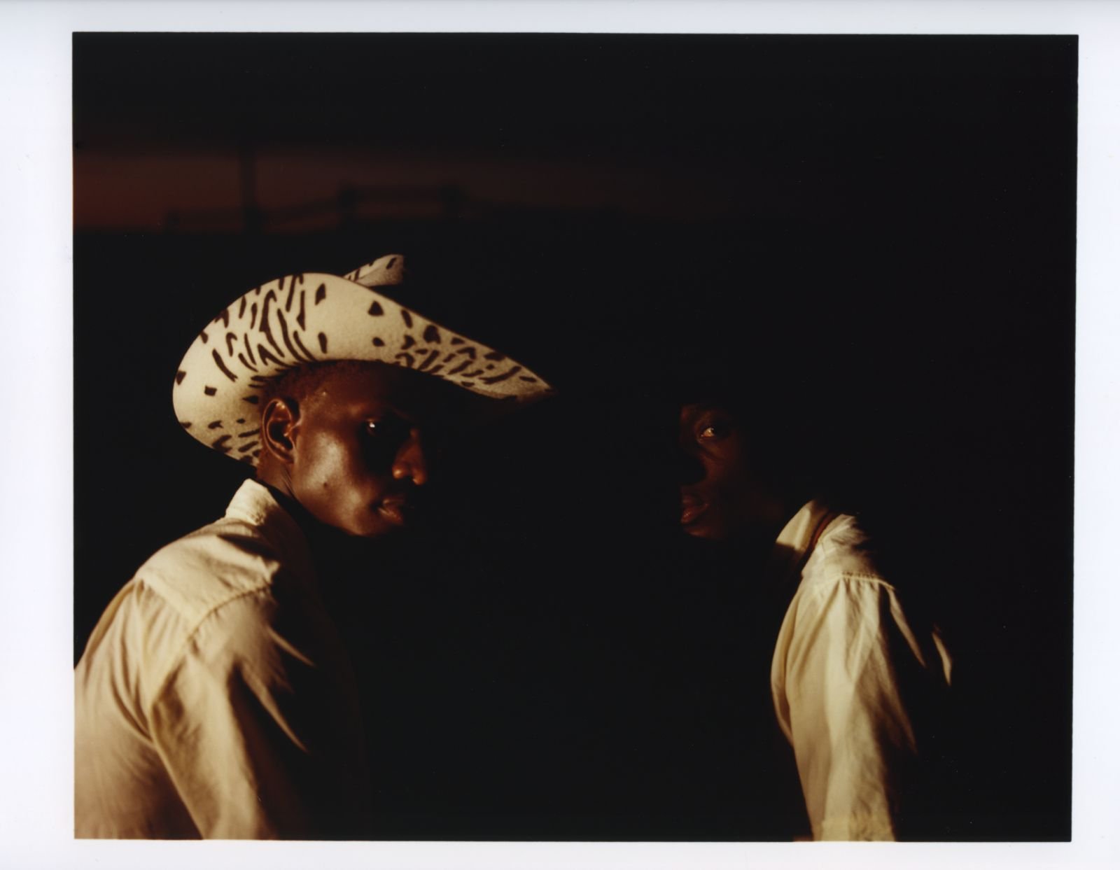 Two young men wearing beige shirts, one with a wide-brimmed, patterned hat, stand in a dark environment, with faint light illuminating their faces.
