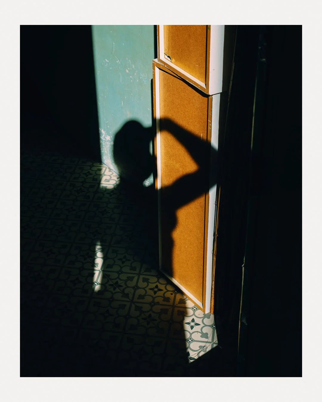 Shadow of a person taking a selfie on a decorative tiled floor, standing near a wooden cabinet and a teal wall with peeling paint.