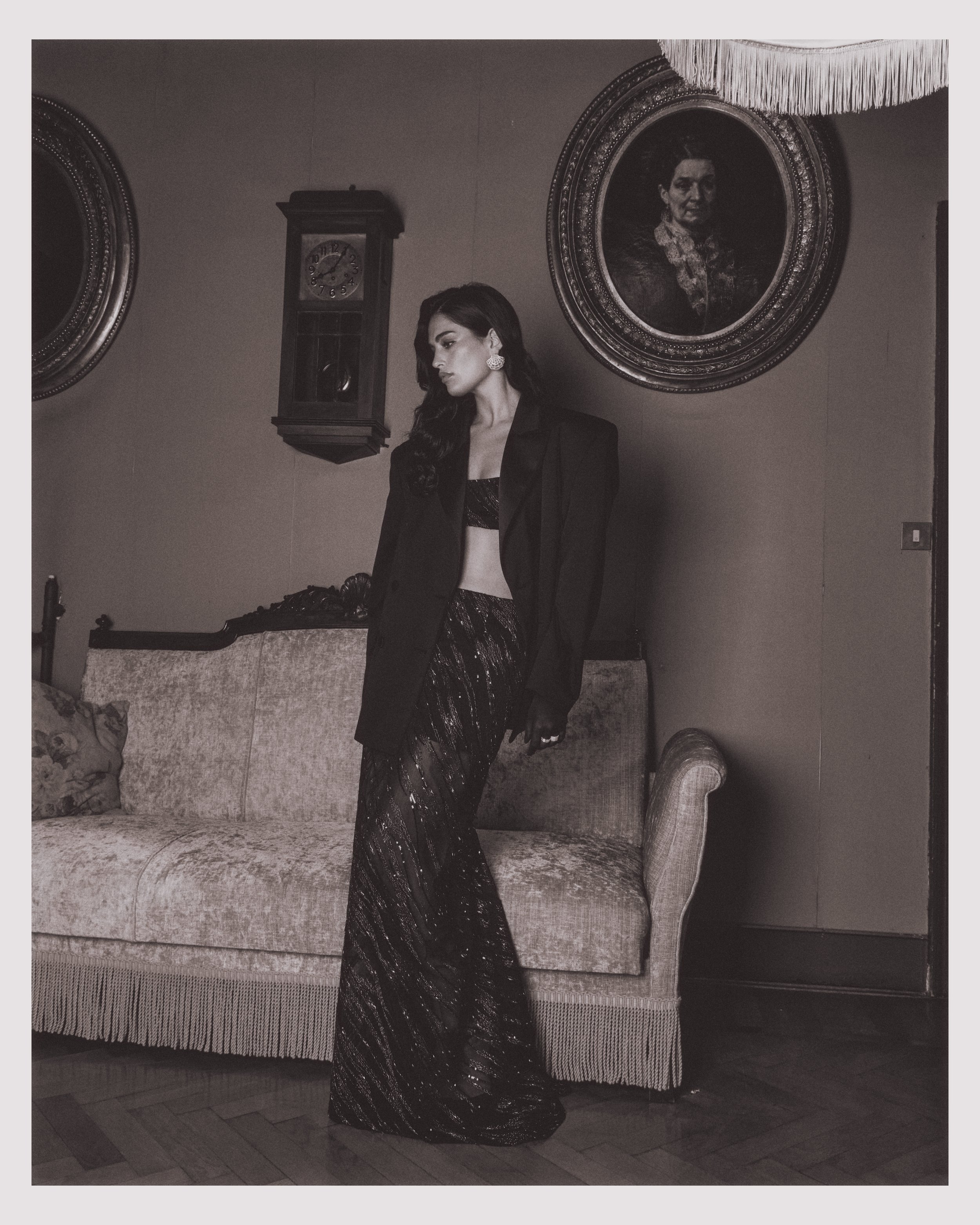 A woman with dark hair in an elegant black gown with a blazer, standing next to a vintage sofa in a room with framed portraits and a wall clock.
