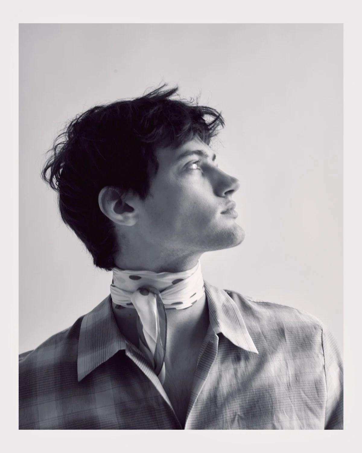 Black and white portrait of a woman with short dark hair, looking upward with a serious expression, wearing a plaid shirt and a polka dot scarf around her neck.