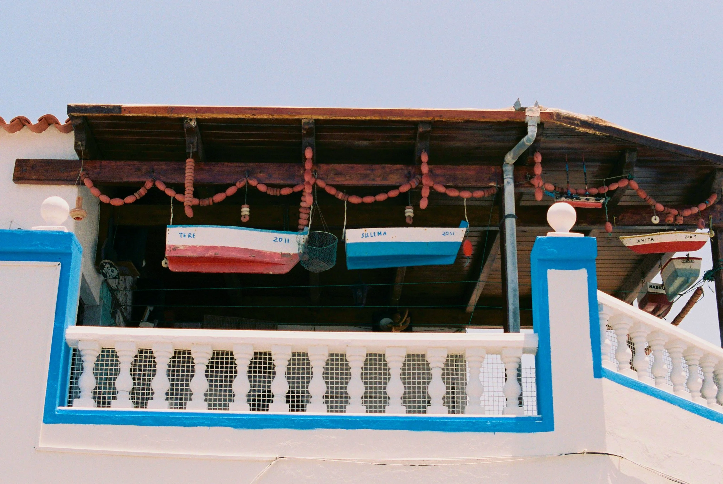 Two small boats hanging from a wooden ceiling above a white balcony with blue trim, decorated with spherical pink and orange beads.