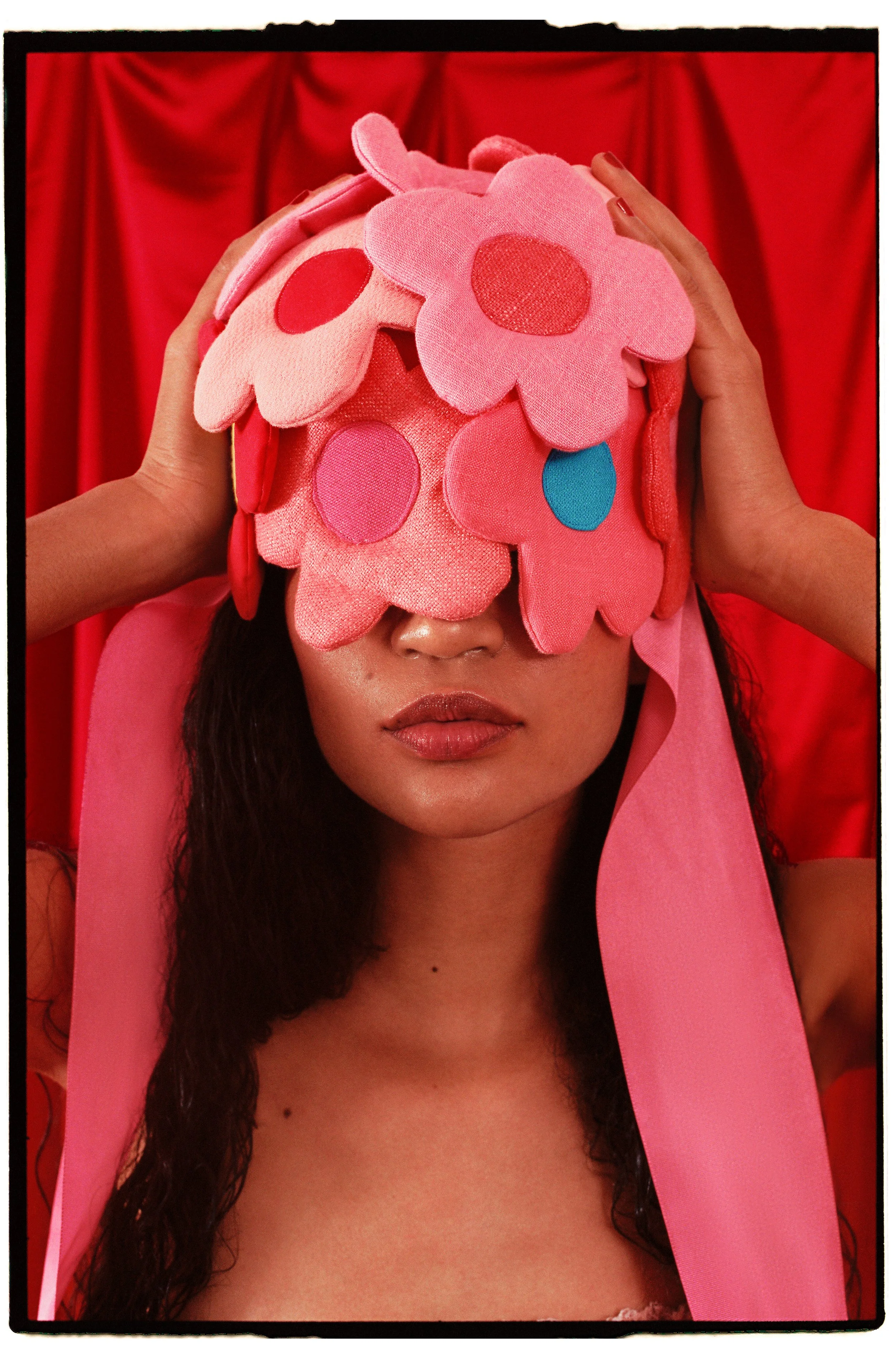 A woman with wet, curly hair wearing a pink flower hat with colorful spots, with a red curtain backdrop.