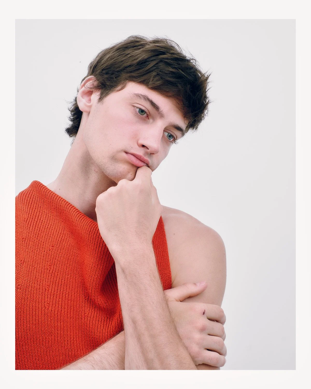 Young man with dark brown hair, light complexion, wearing an orange sleeveless knitted top, resting his chin on his hand, in front of a plain white background.