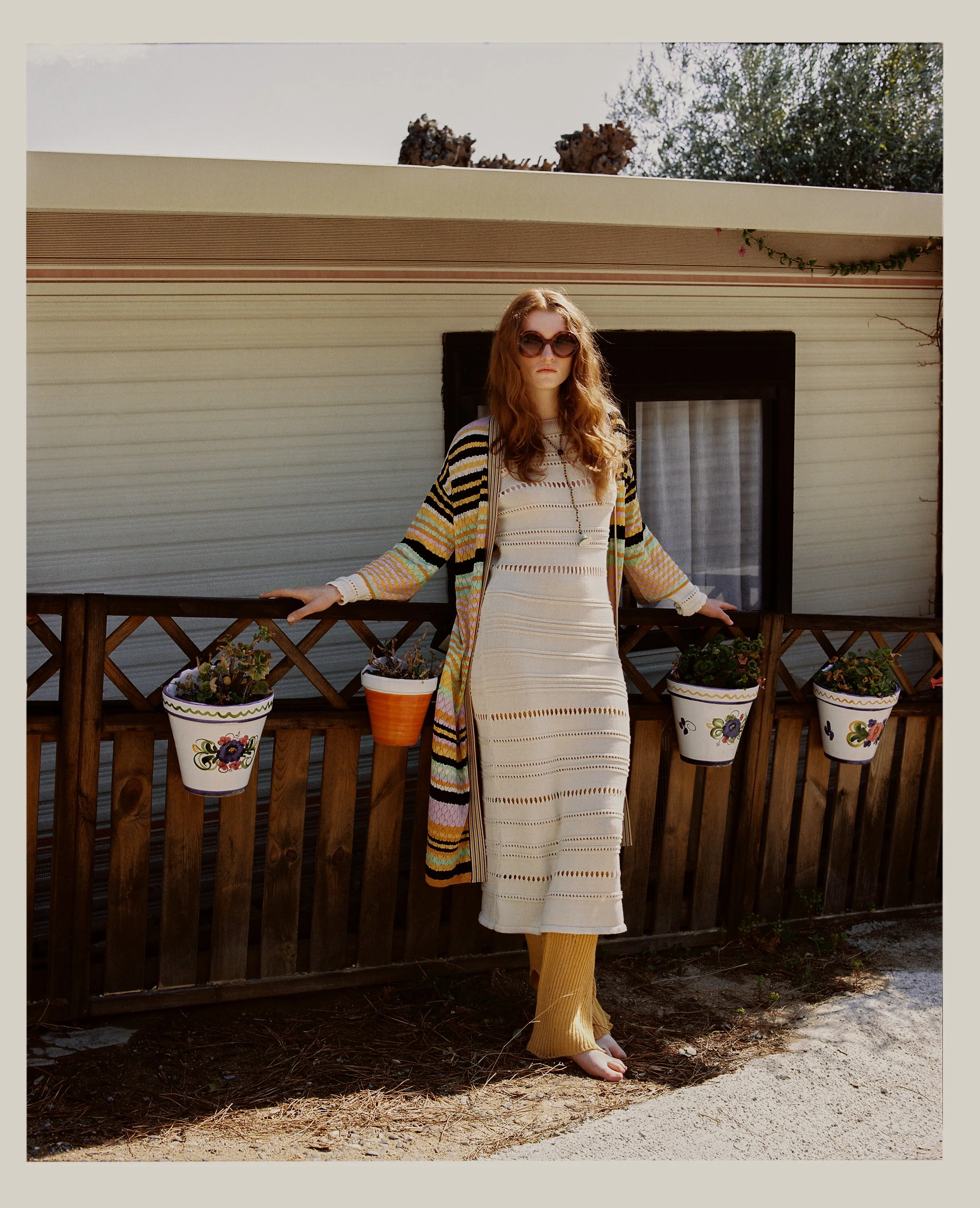 Woman with long wavy hair wearing large sunglasses, a cream-colored dress with horizontal cutout patterns, a multicolored striped cardigan, and yellow knit pants, standing barefoot outdoors by a wooden fence with flower pots, in front of a mobile hom