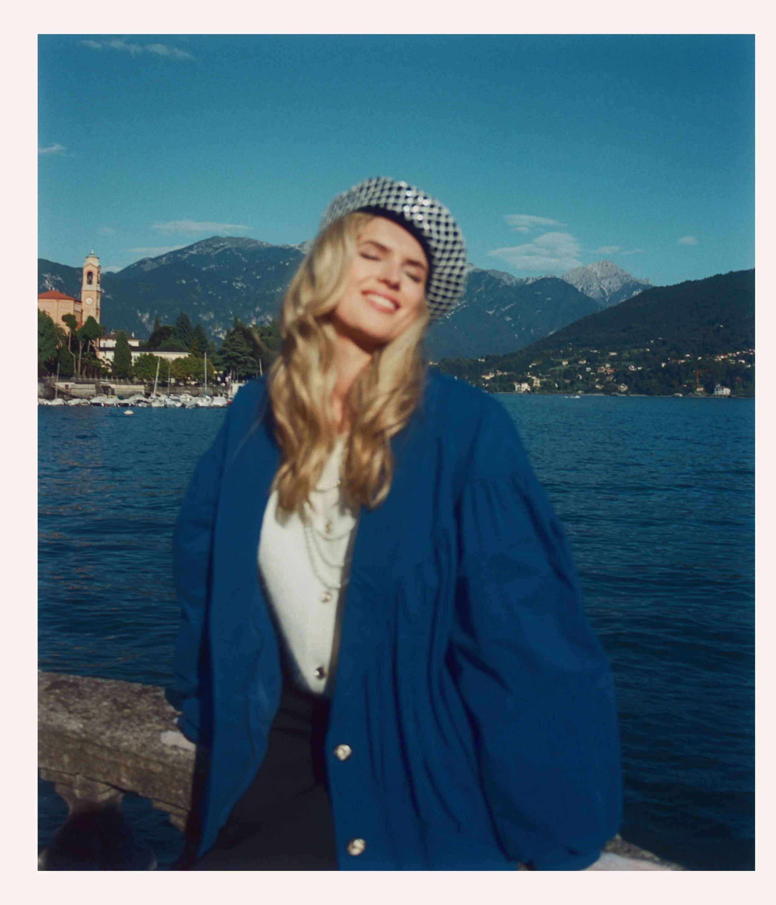 A woman with long blonde hair smiling with her eyes closed, wearing a blue jacket and a hat with a checkered pattern, standing by a lake with mountains and a small town in the background.