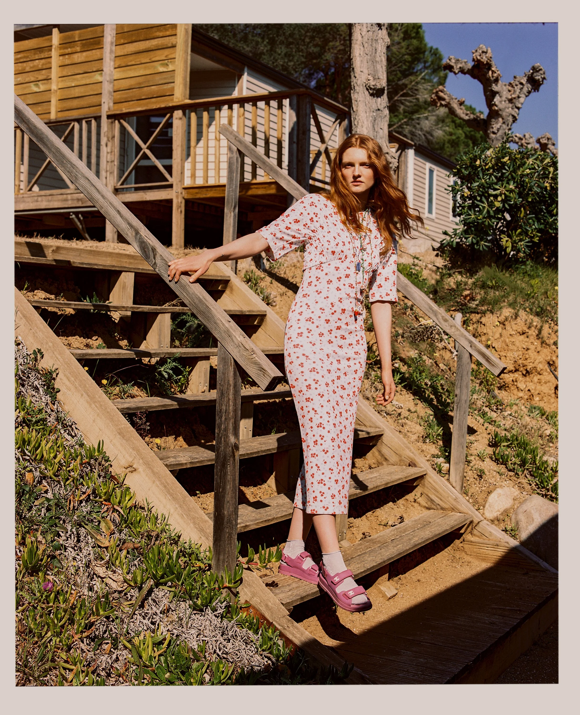 A young woman with red hair wearing a white dress with a red floral pattern, pink sandals, and white socks, stands on wooden stairs outdoors near a house with wooden siding, trees, and shrubs in the background.