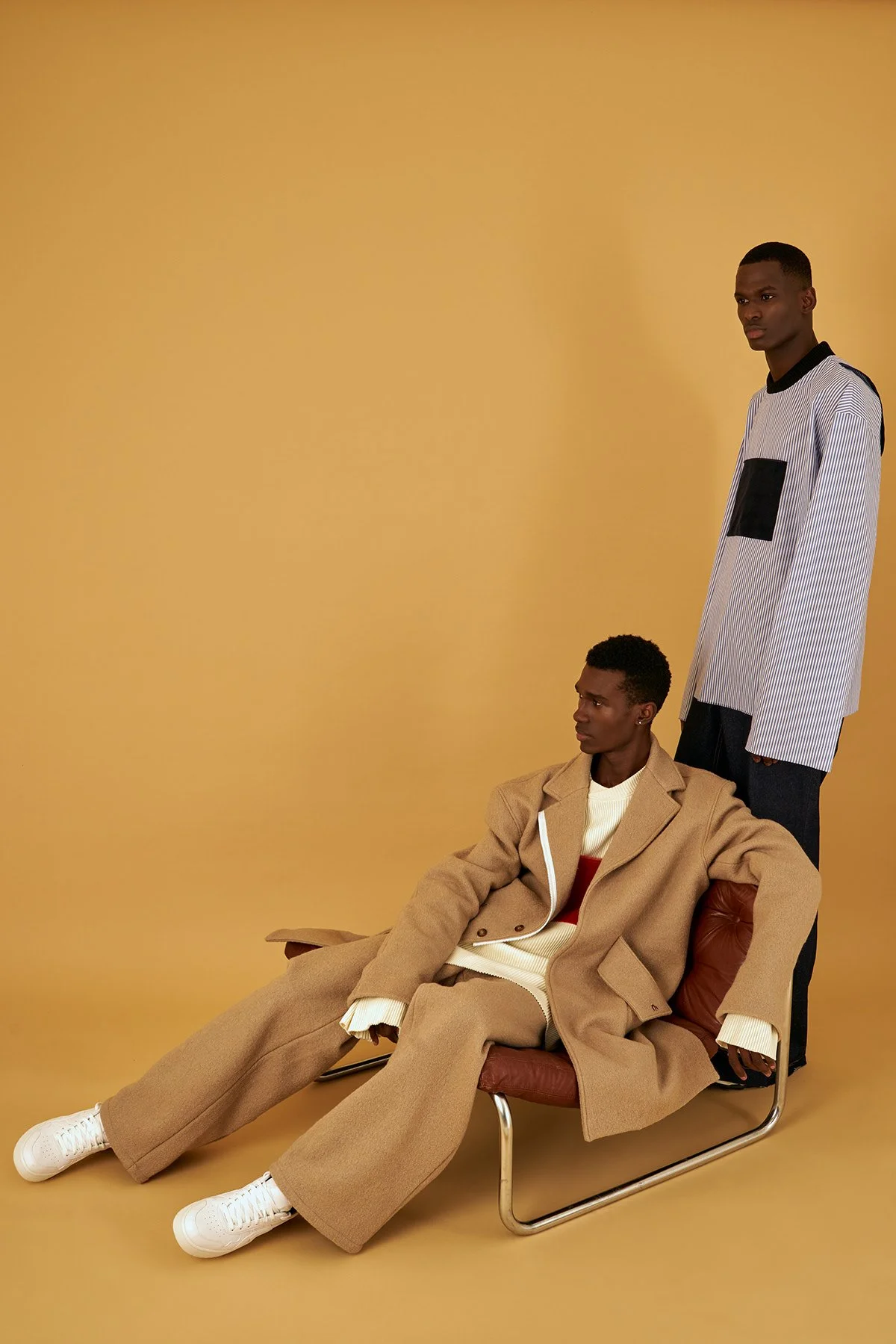 Two men wearing stylish clothing against a beige background. One is sitting on a brown chair, wearing a beige oversized coat, cream sweater, and white sneakers. The other is standing, dressed in a striped white and black shirt with a black pocket det