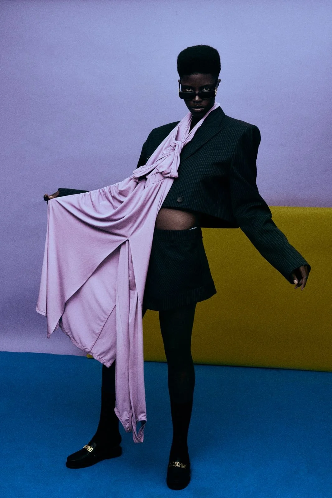 A person with dark skin and a high flat-top hairstyle, wearing black sunglasses, a black pinstripe blazer, matching shorts, black tights, black shoes with gold elements, and a large light pink satin scarf draped over their shoulder against a multicol
