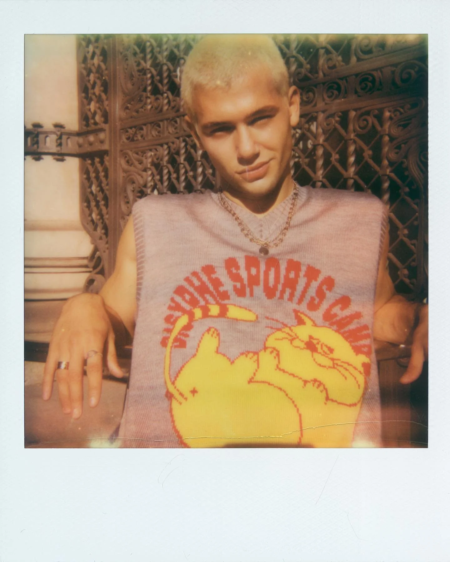 A person with short platinum blonde hair sitting in front of a decorative metal gate, wearing a sleeveless gray top with a yellow graphic of a cat and red text, layered necklaces, rings, and making a casual pose with hands resting on the arms of a ch