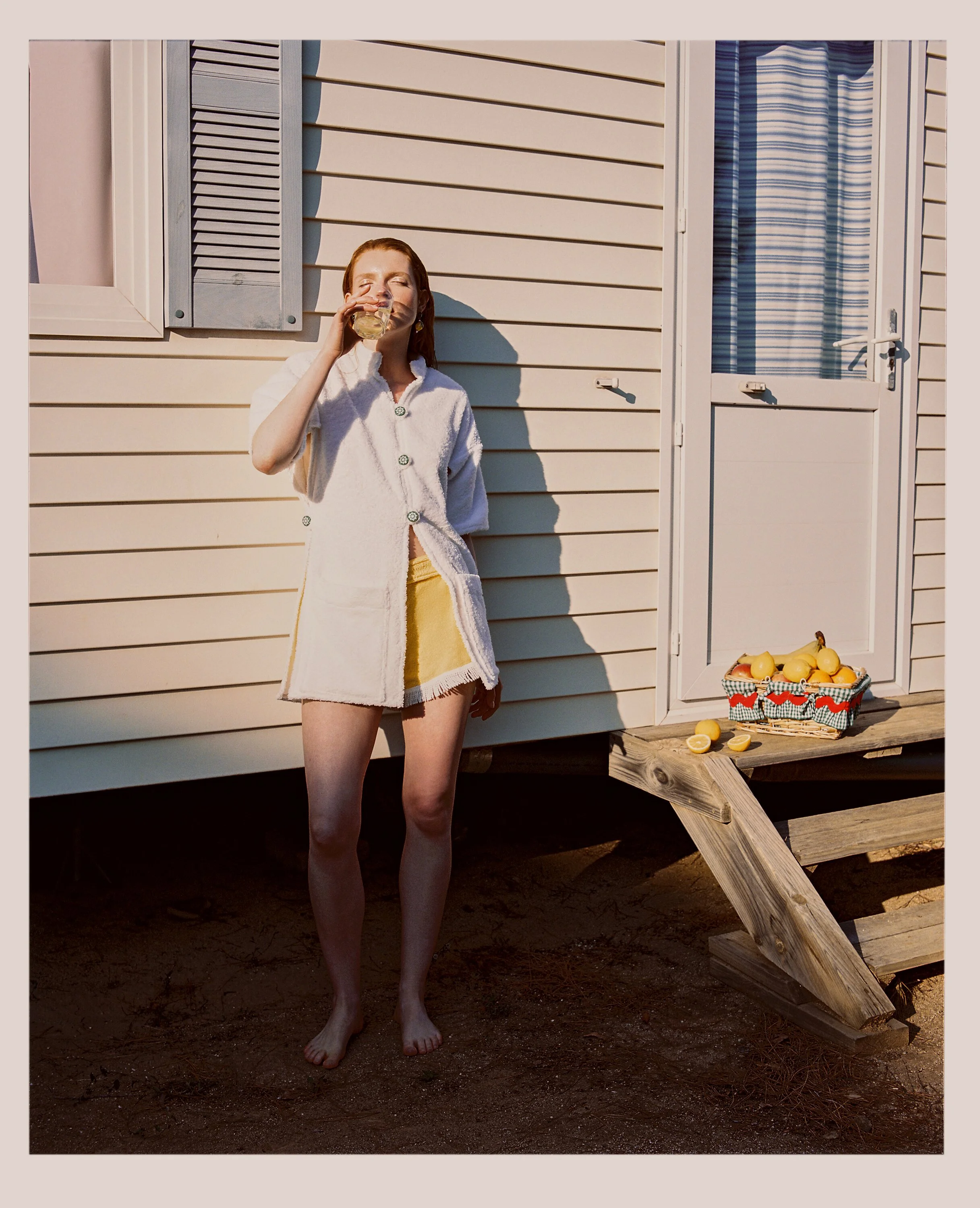 a woman with red hair wearing a white bathrobe and yellow shorts, standing outside a beige house, drinking a beverage, with a basket of lemons on a small wooden staircase nearby.